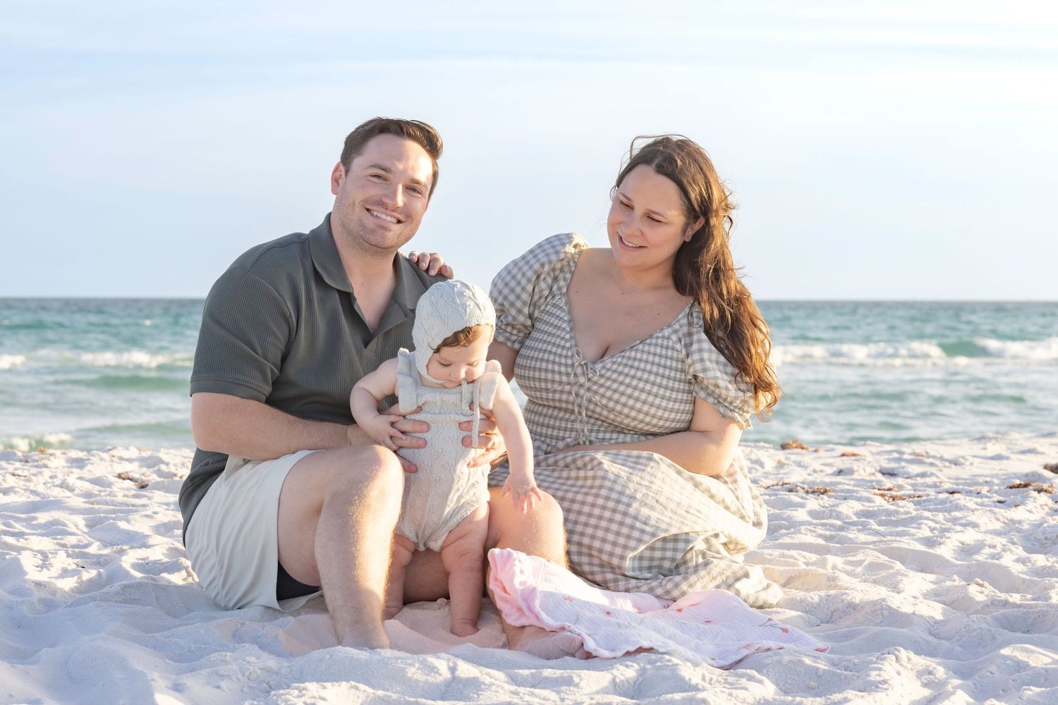 Family of three smiling on a sandy beach, parents sitting with their baby on a sunny day near the ocean.