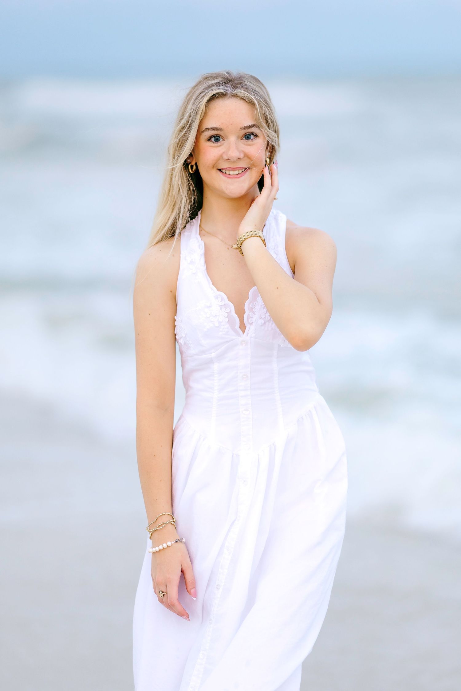 Young blonde woman in white halter dress smiling at beach with ocean waves in background.