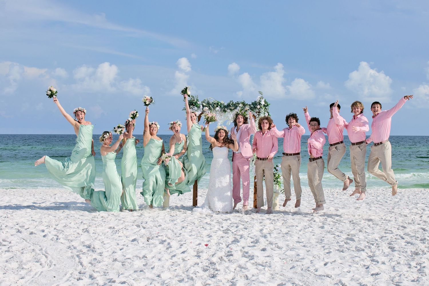 Wedding party jumping joyfully on a sunny beach, bridesmaids in mint green and groomsmen in pink shirts.