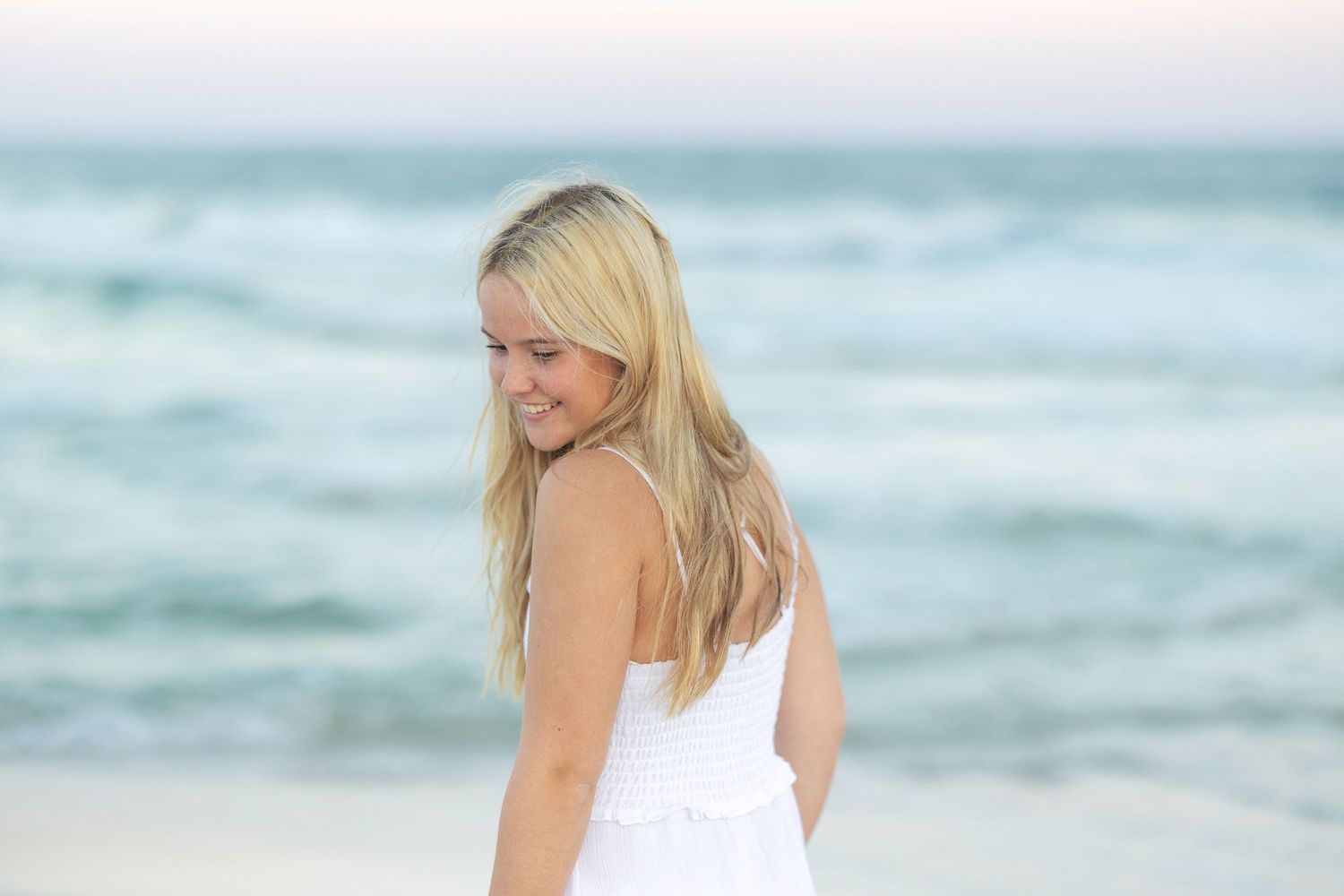 Young blonde woman in white dress smiling over her shoulder on a sandy beach with ocean waves in the background.