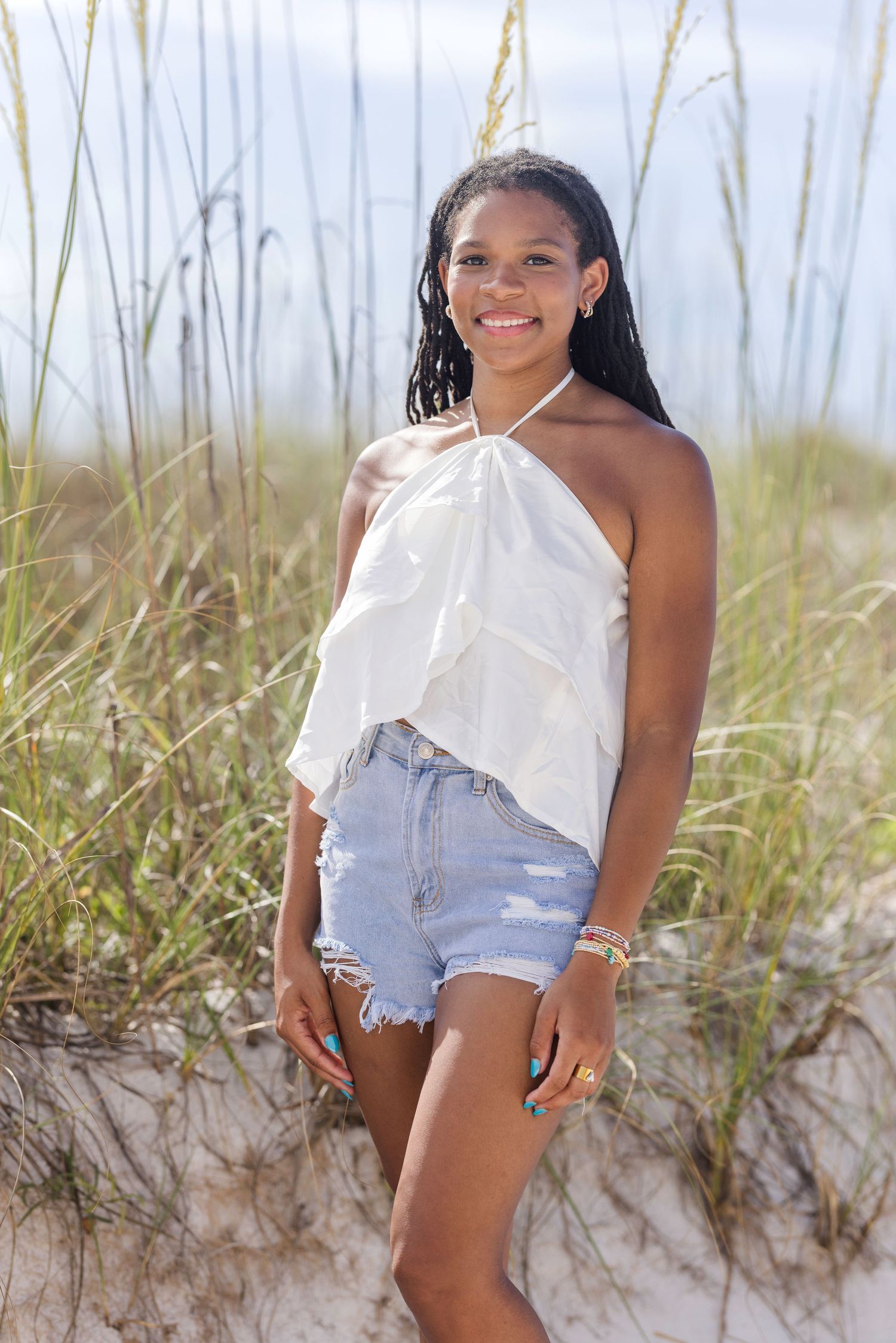 Young Black woman smiling on sandy beach dunes, wearing white halter top and denim cutoff shorts, with sea oats behind her.