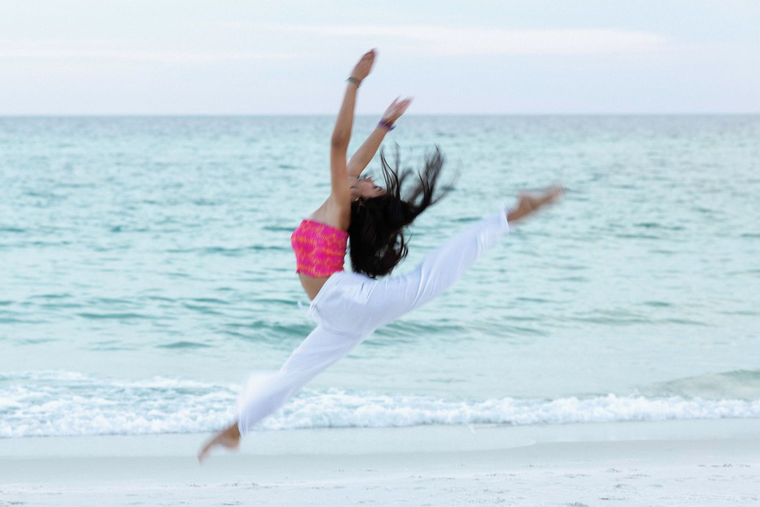 A dancer leaps gracefully across a pristine beach with turquoise ocean waters in the background.