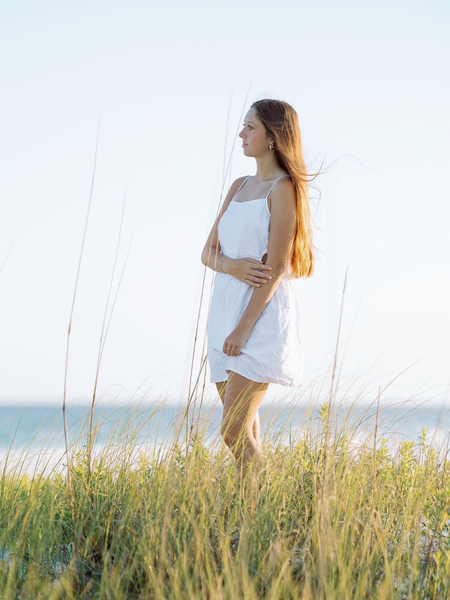 Woman in flowing white dress stands among tall beach grass during golden hour photoshoot.
