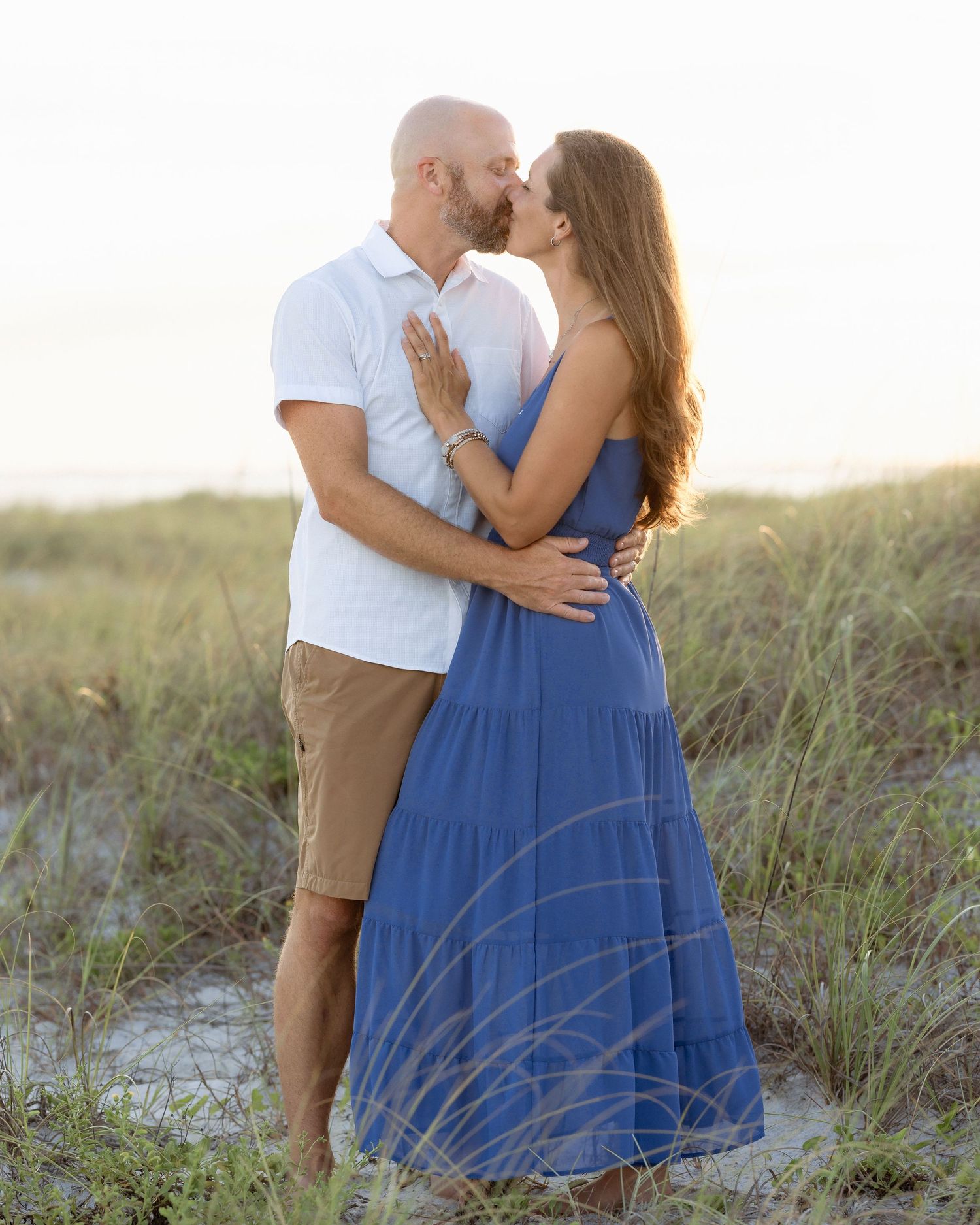 Couple sharing a romantic kiss on sandy beach dunes at sunset, woman in blue maxi dress, man in white shirt and khaki shorts.