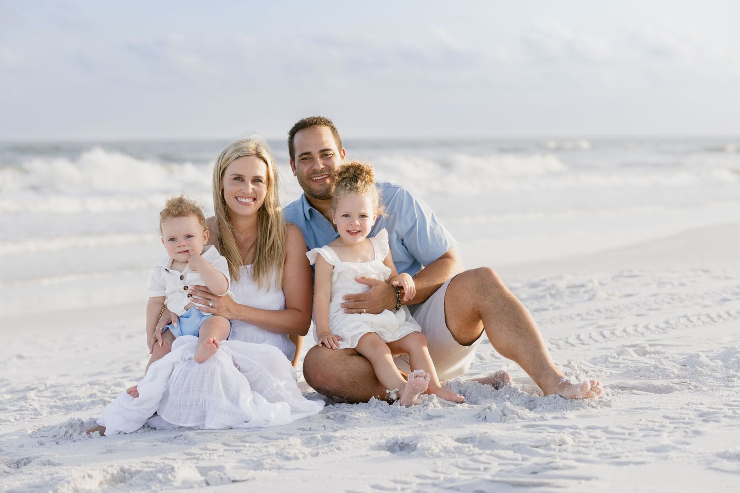 Happy family of four posing on a sandy beach, parents holding two young children with ocean waves in the background.
