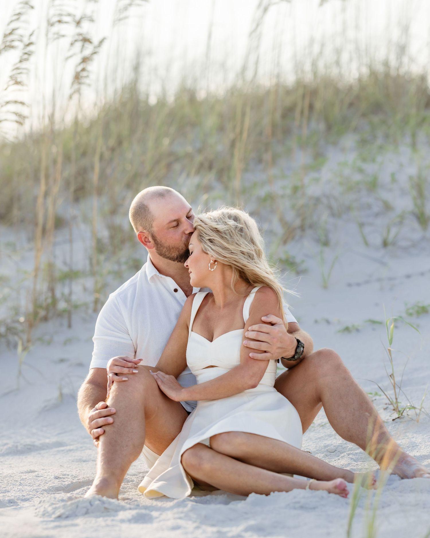 Couple sitting on beach sand dunes, man kissing woman's cheek, both wearing white, with sea oats in background.