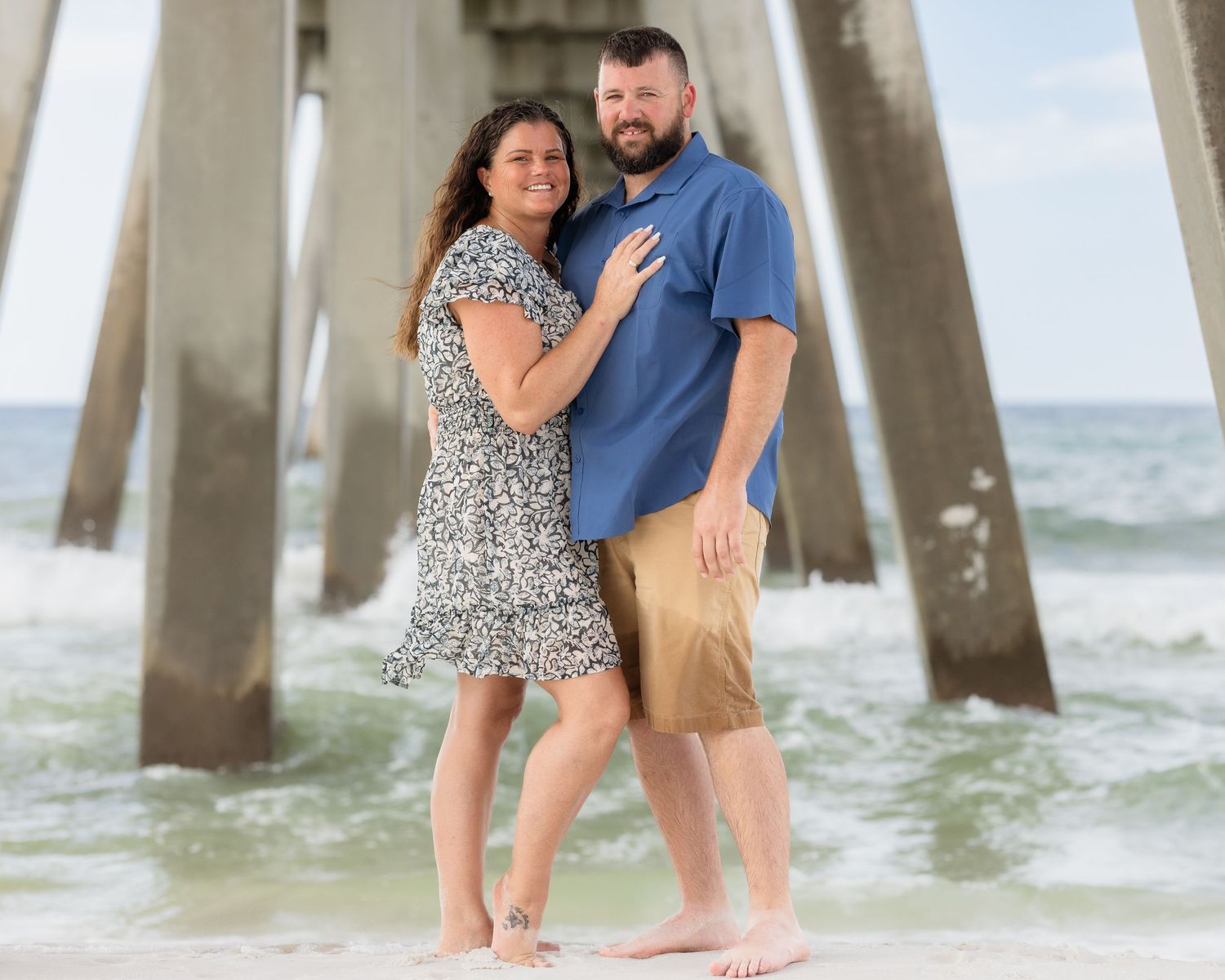 Couple embraces barefoot on beach under pier with ocean waves in the background.