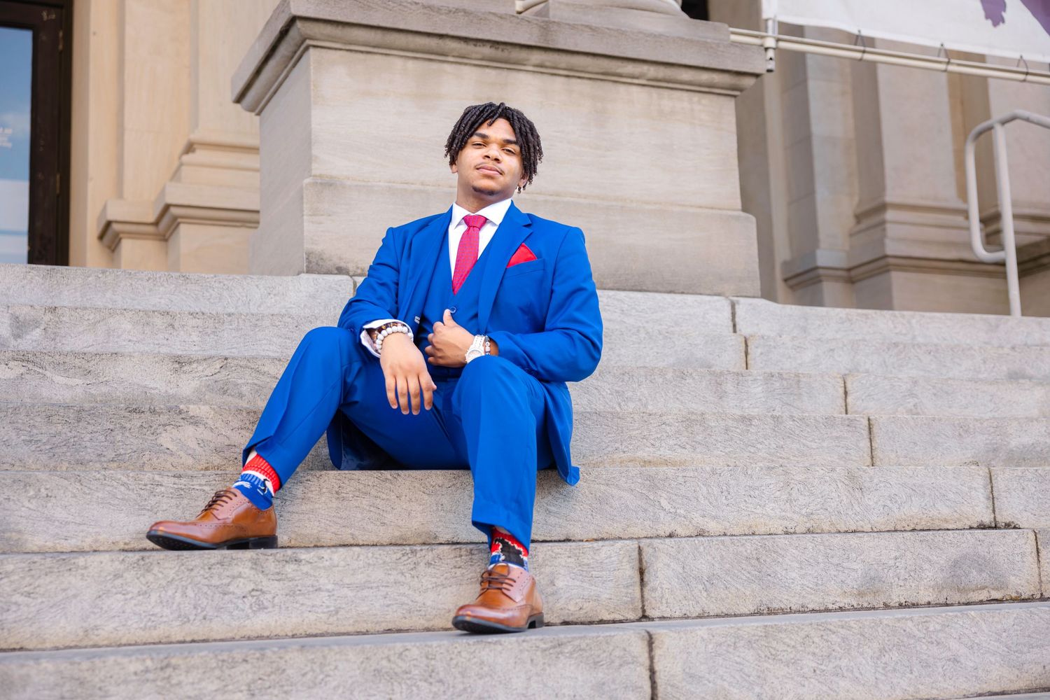 Man in blue suit with red tie sitting on stone steps, wearing brown dress shoes and colorful socks.