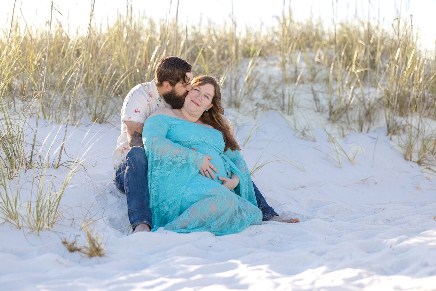 Couple sitting on white sand beach dunes, pregnant woman in teal lace gown leaning against partner during maternity photo session.