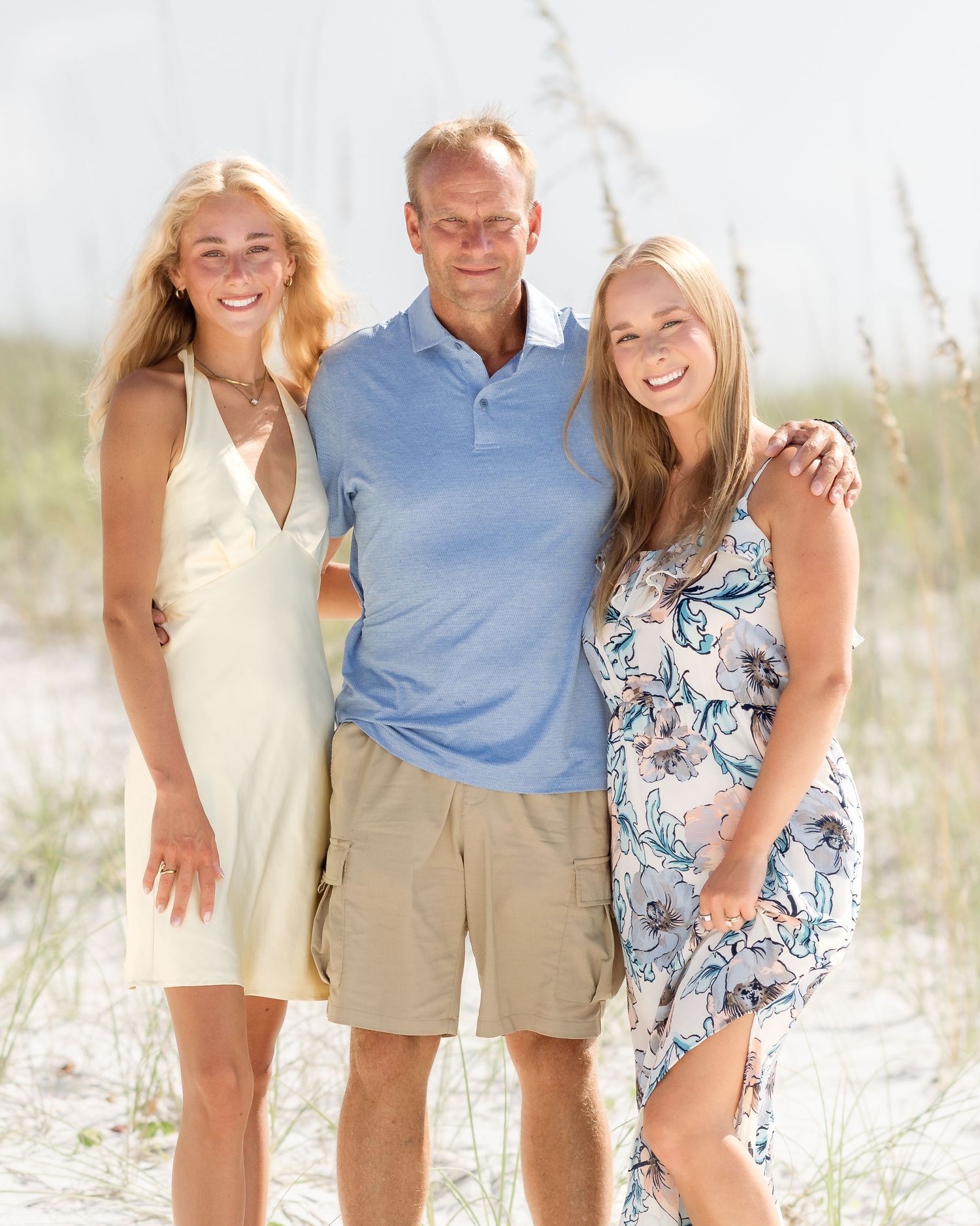 A man and two blonde women smile together on a sunny beach with white sand dunes and sea oats in the background.
