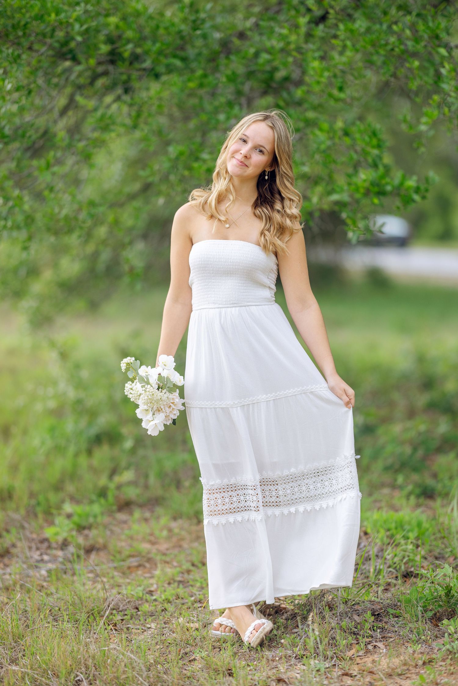 White summer maxi dress with lace details is styled with sandals and white floral bouquet in an outdoor nature setting.