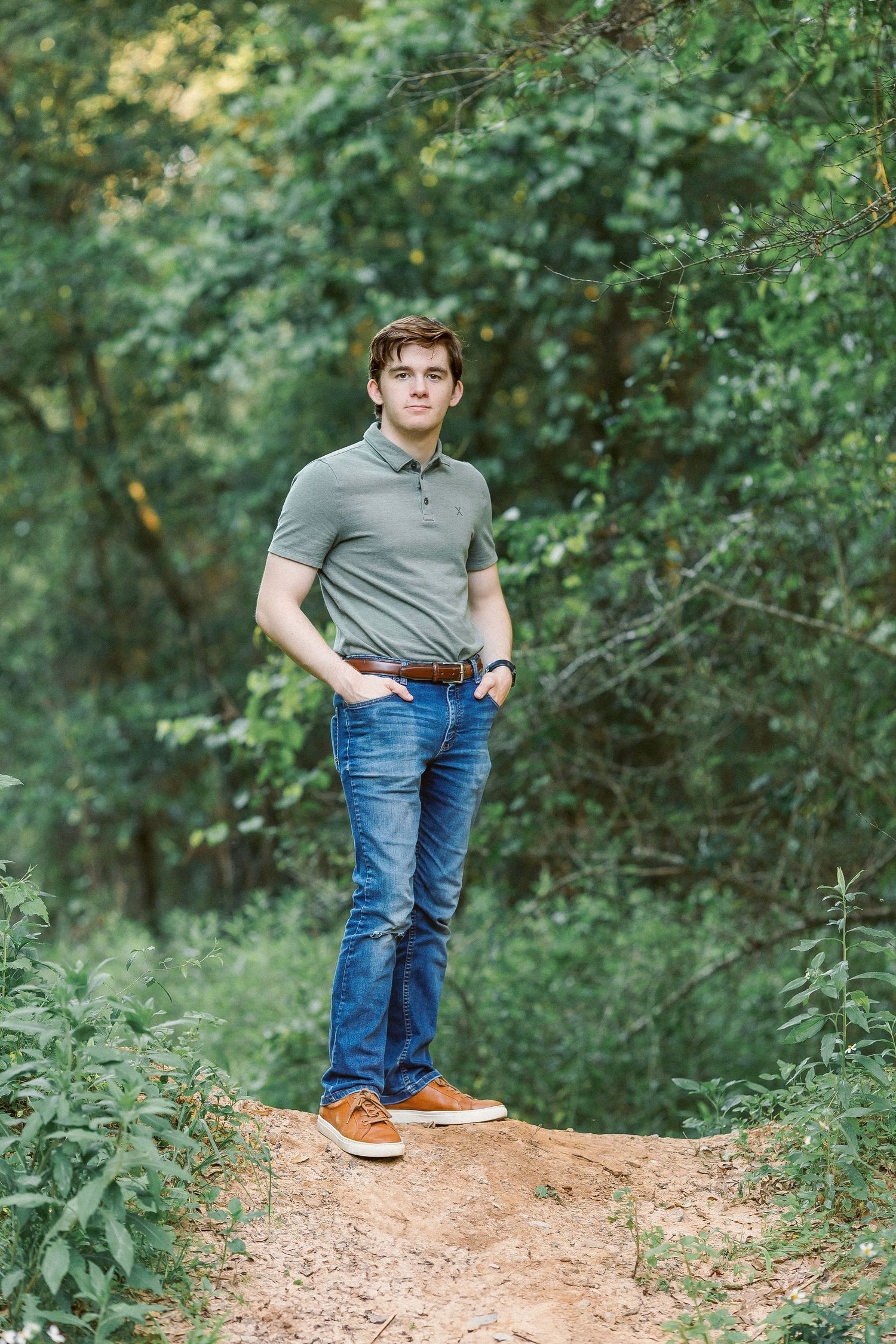 Portrait photograph in a nature setting with casual attire and brown shoes standing on a dirt path amid green foliage.