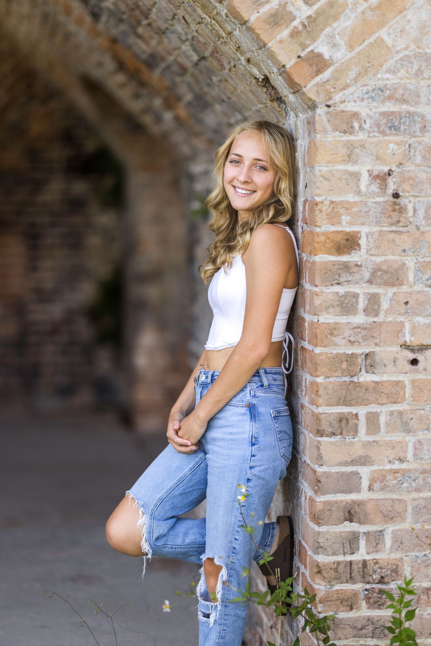 A young woman in ripped jeans and a white top poses casually against a rustic brick archway.