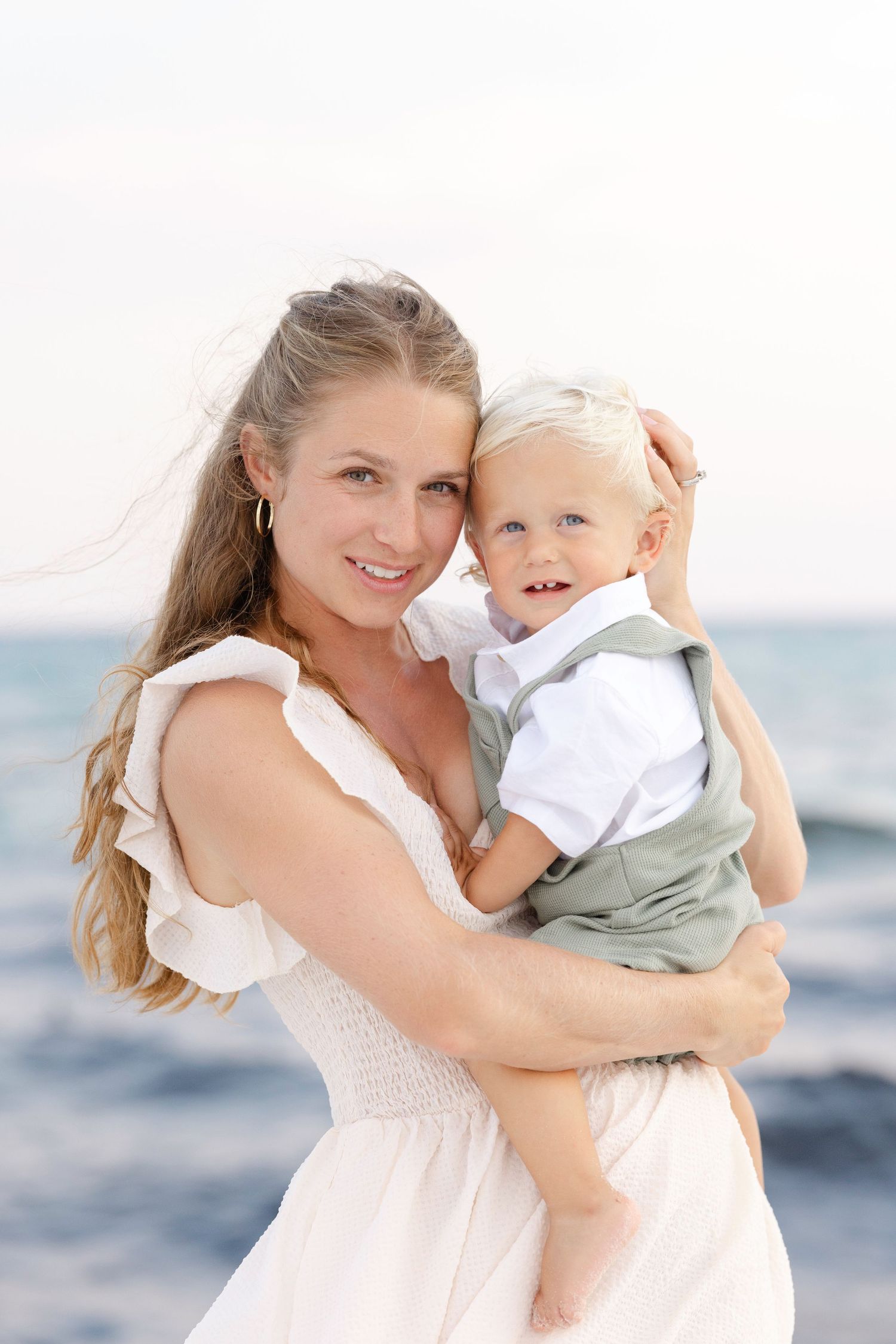 A smiling woman in a white dress holds a happy toddler boy near the ocean on a sunny day.