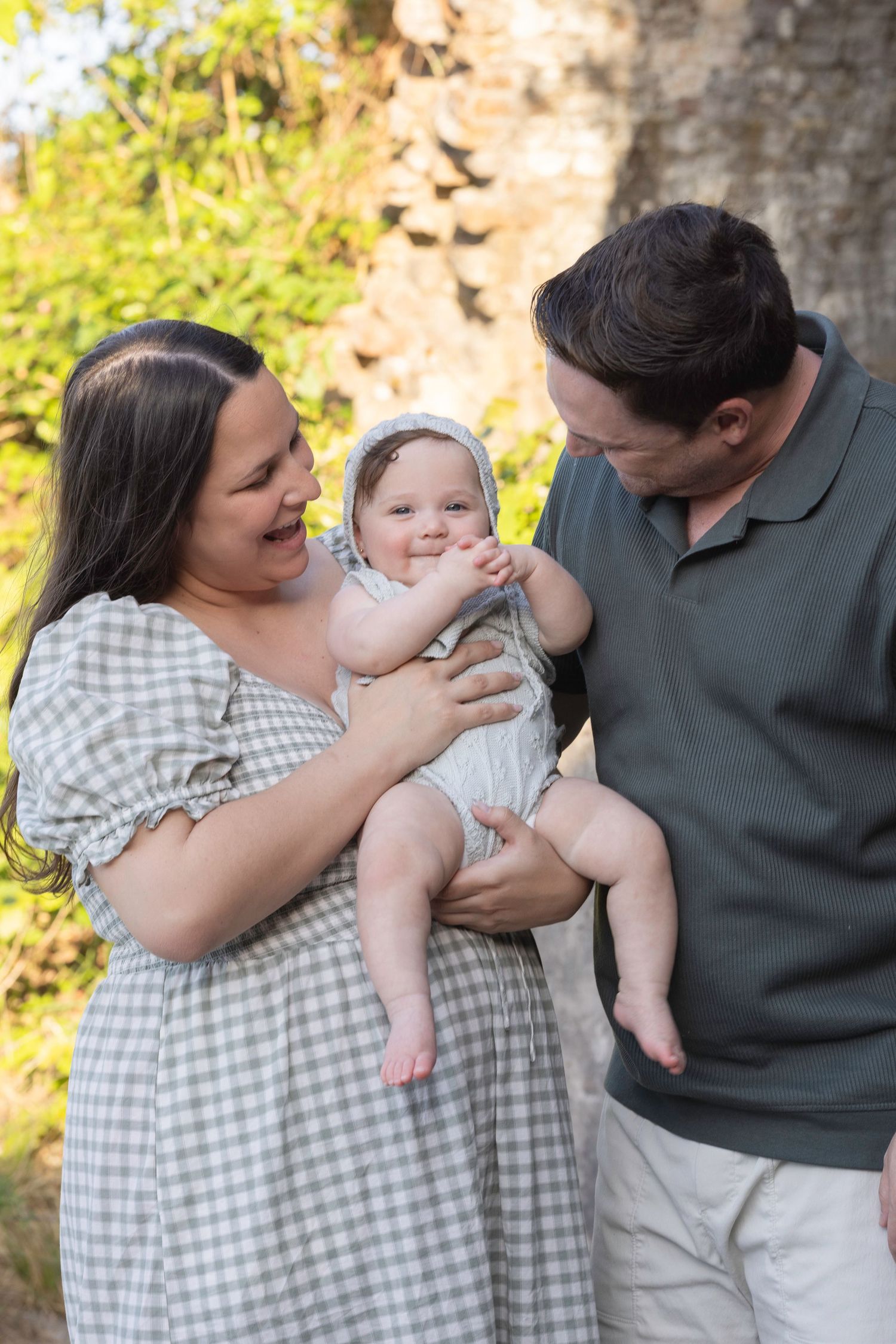 Happy family with mother in plaid dress and father in green polo holding smiling baby outdoors near tree.