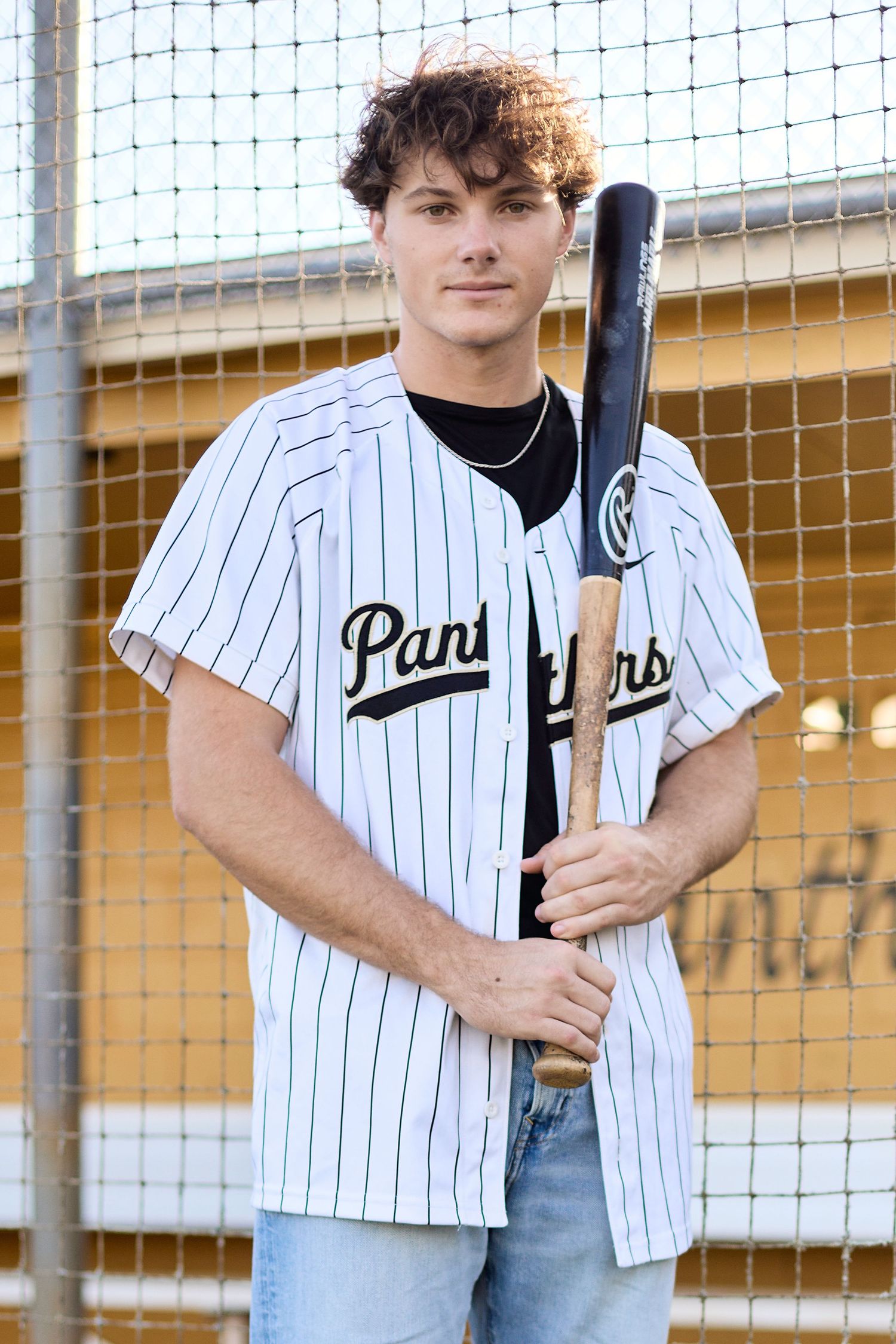 Young man in Panthers baseball jersey holding a bat, posing near a chain-link fence outdoors.