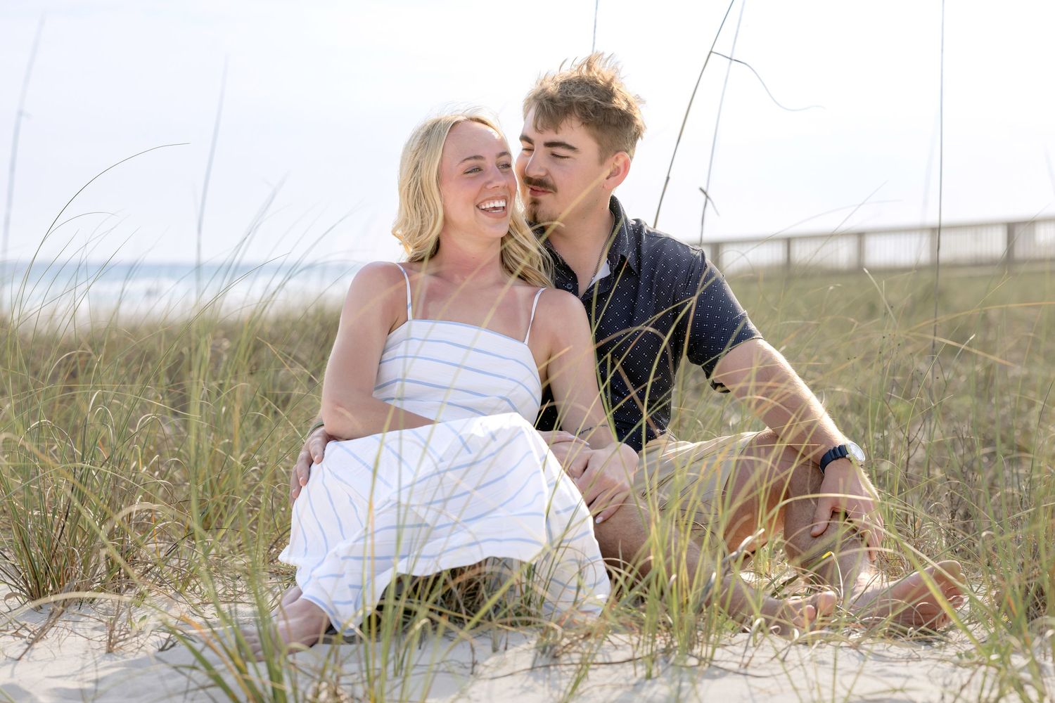 A happy couple sits together on sandy beach dunes, sharing a romantic moment with ocean and wooden fence in background.
