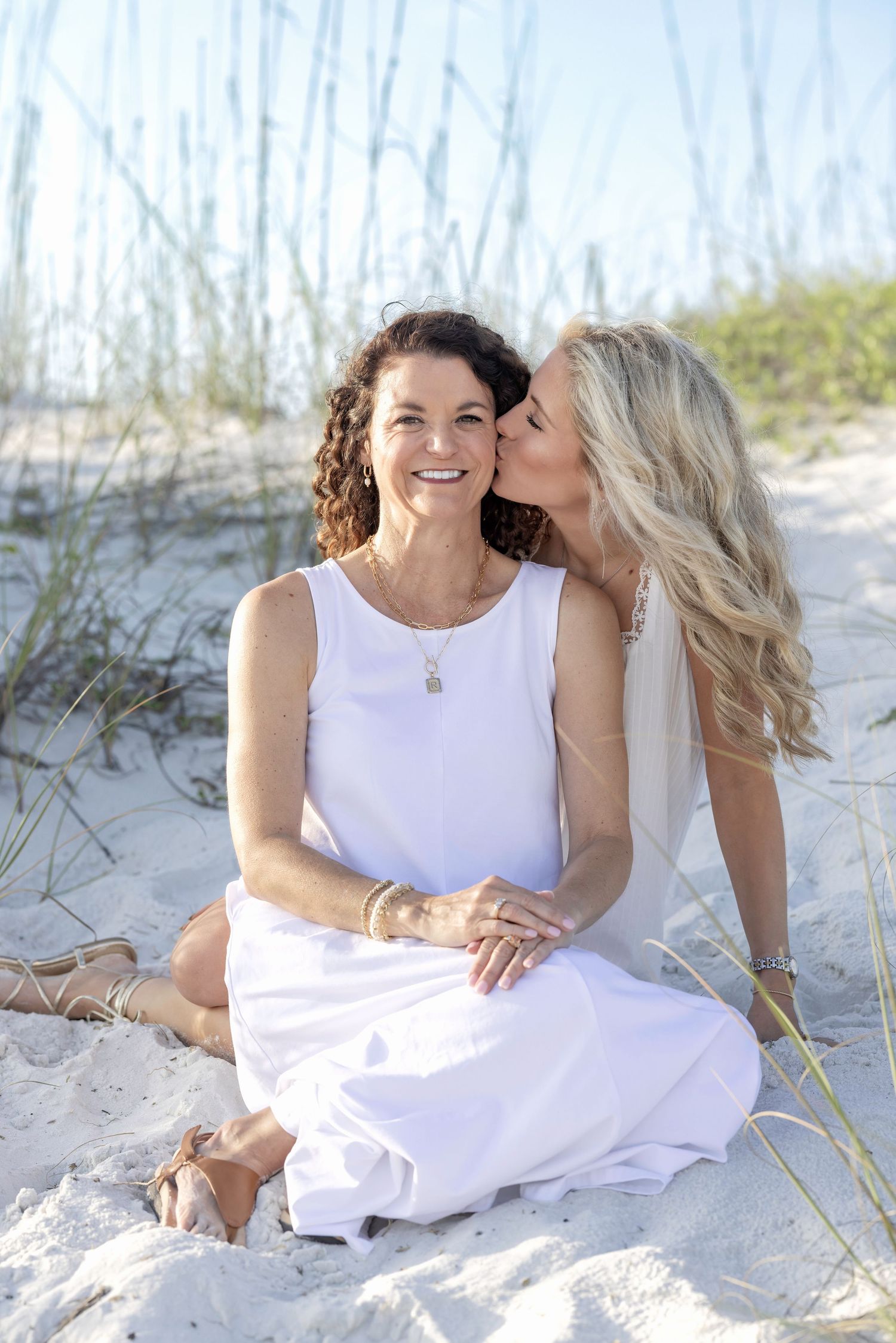 Two women in white dresses sit on beach sand dunes, one kissing the other on the cheek near sea oats.