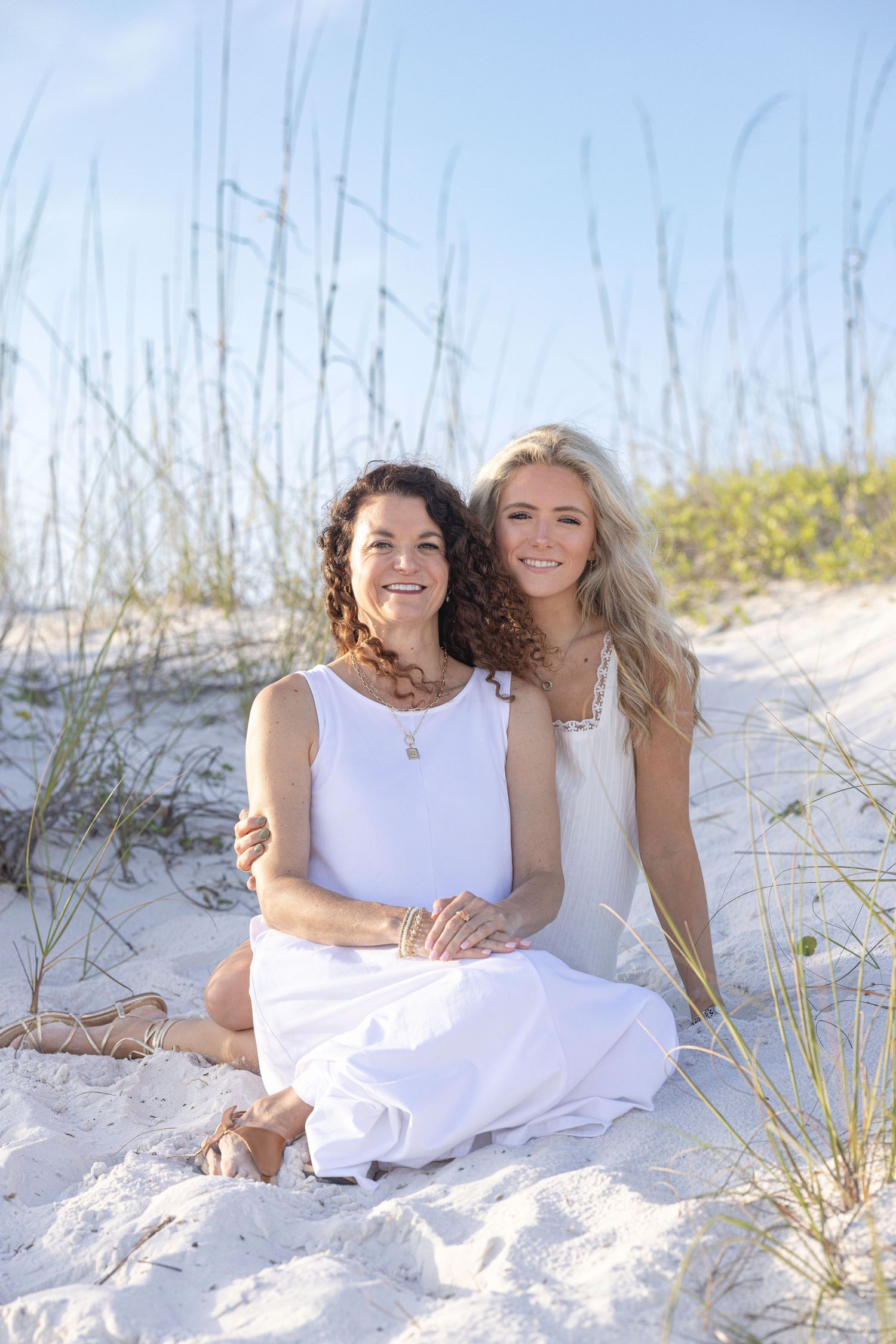 Two women in white dresses sit together on sandy beach dunes, smiling warmly with sea oats in the background.
