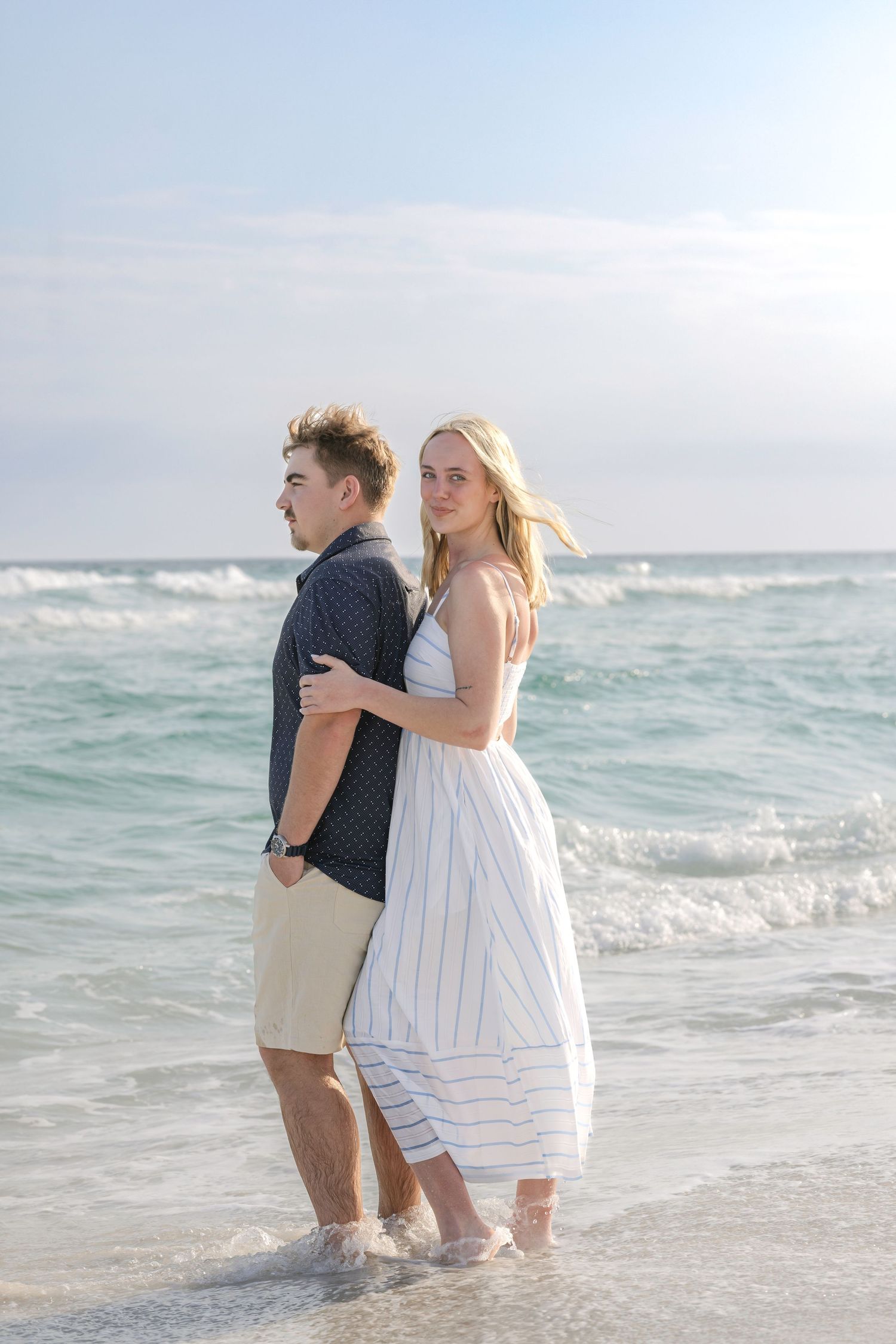 A young couple stands together on a sunny beach, the woman in a white dress embracing the man as waves roll in behind them.