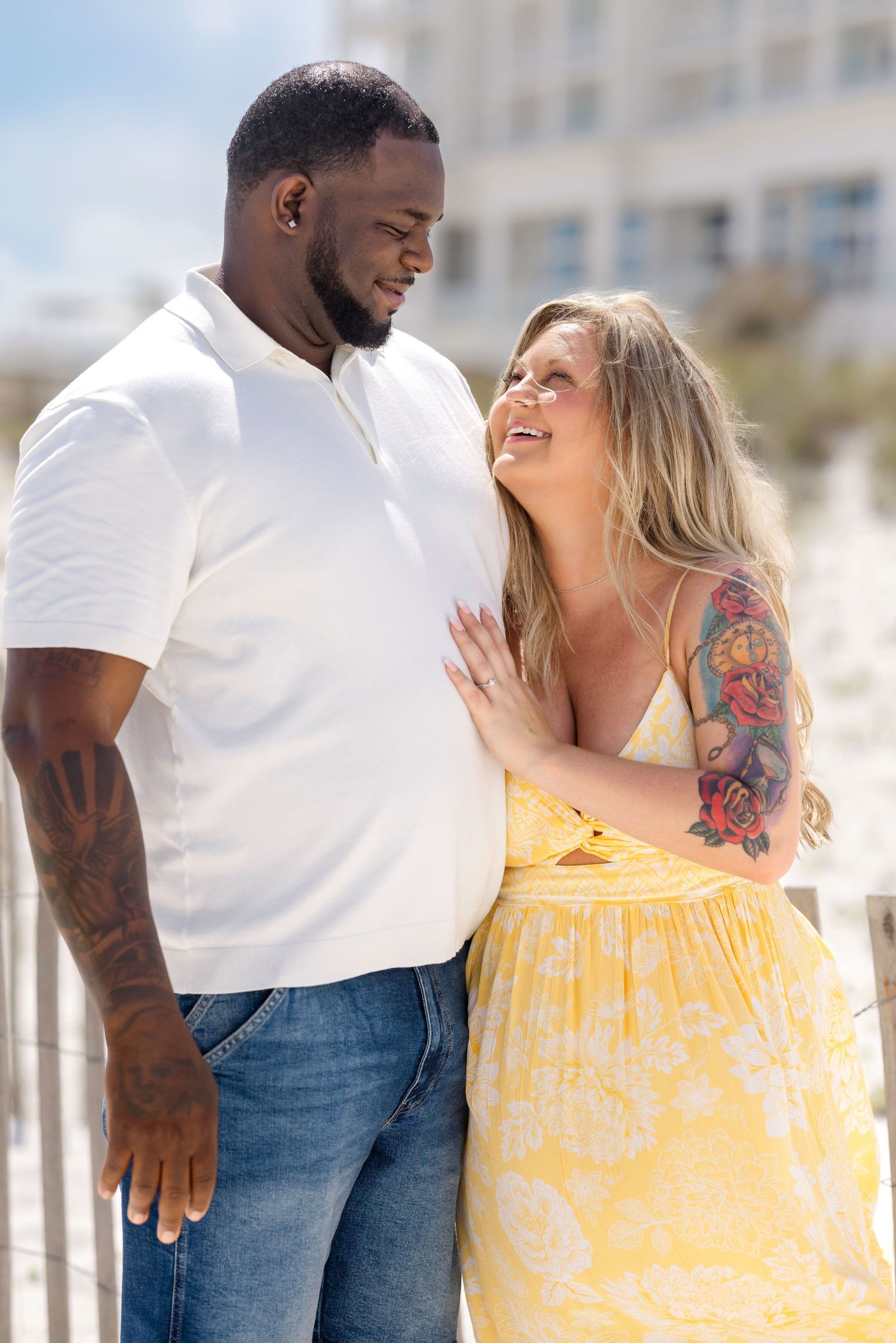 Interracial couple embracing and smiling at each other near a beach, woman in yellow dress with tattoos, man in white polo.