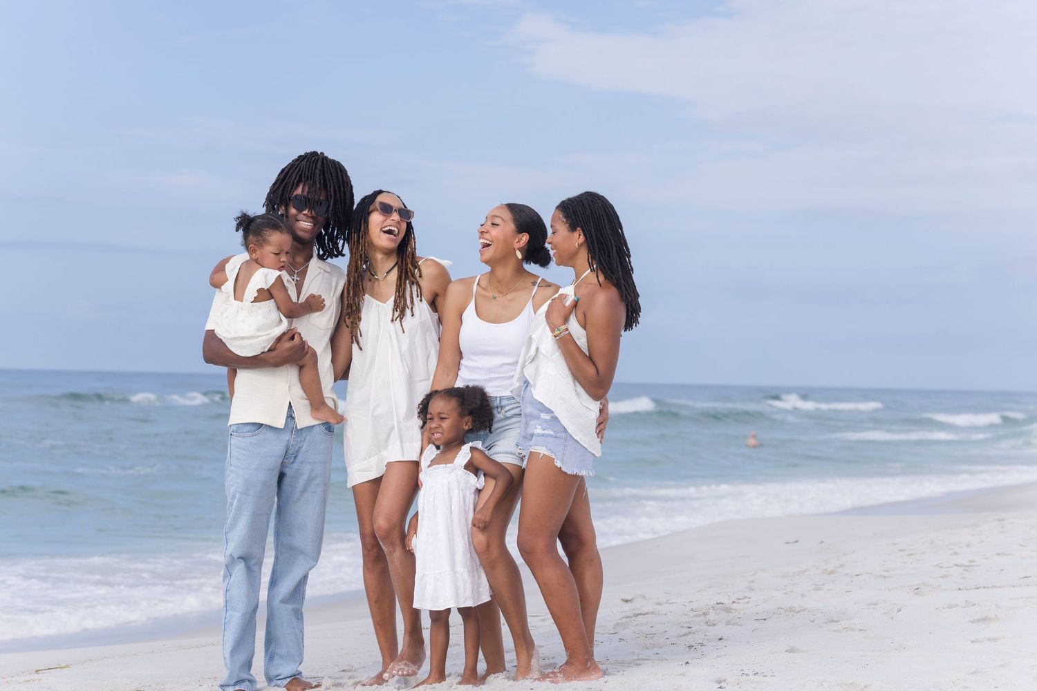 A joyful multiracial family of six smiling together on a sunny beach with ocean waves in the background.