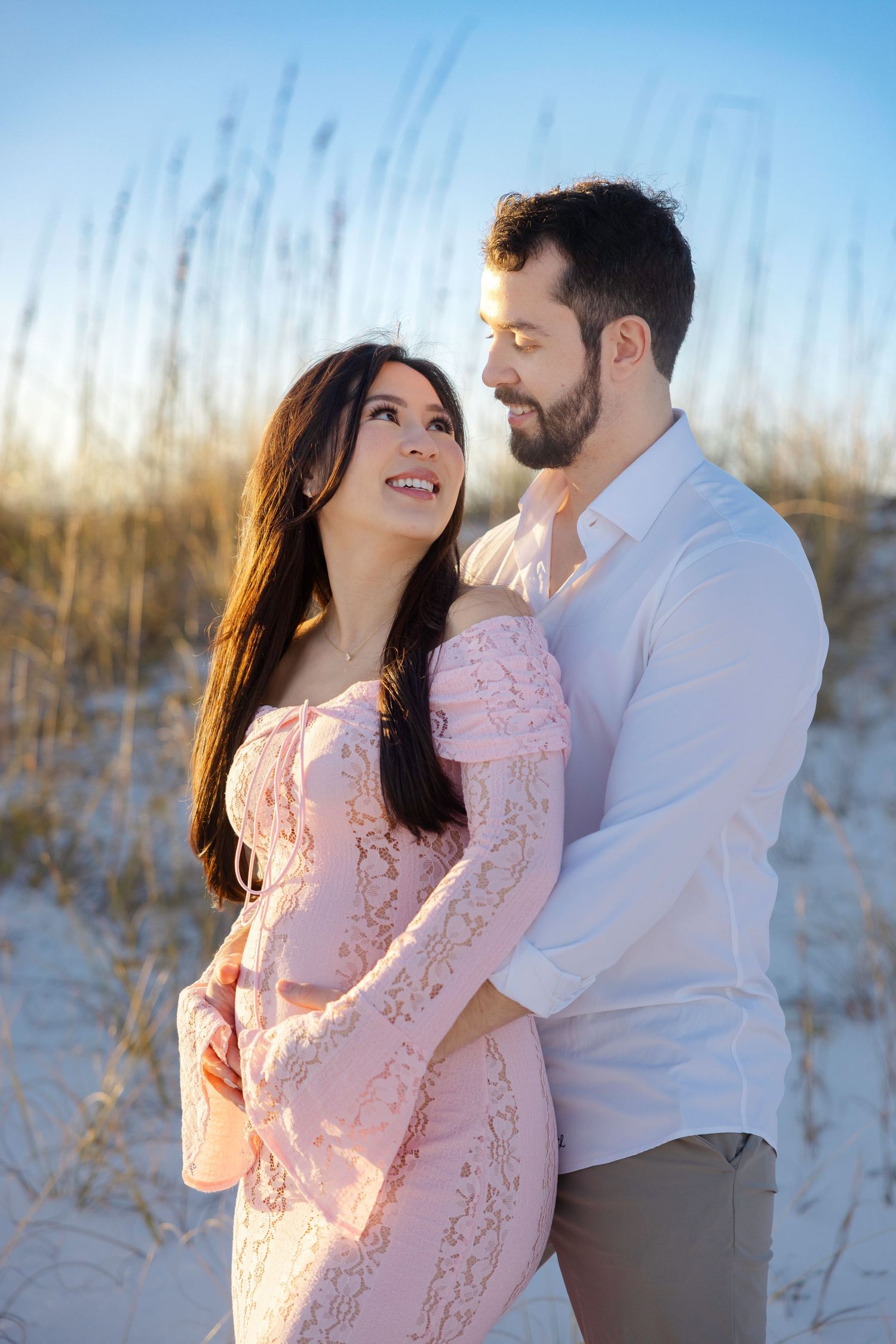 A couple embraces on a sandy beach at sunset, woman in pink lace dress smiling up at bearded man in white shirt.