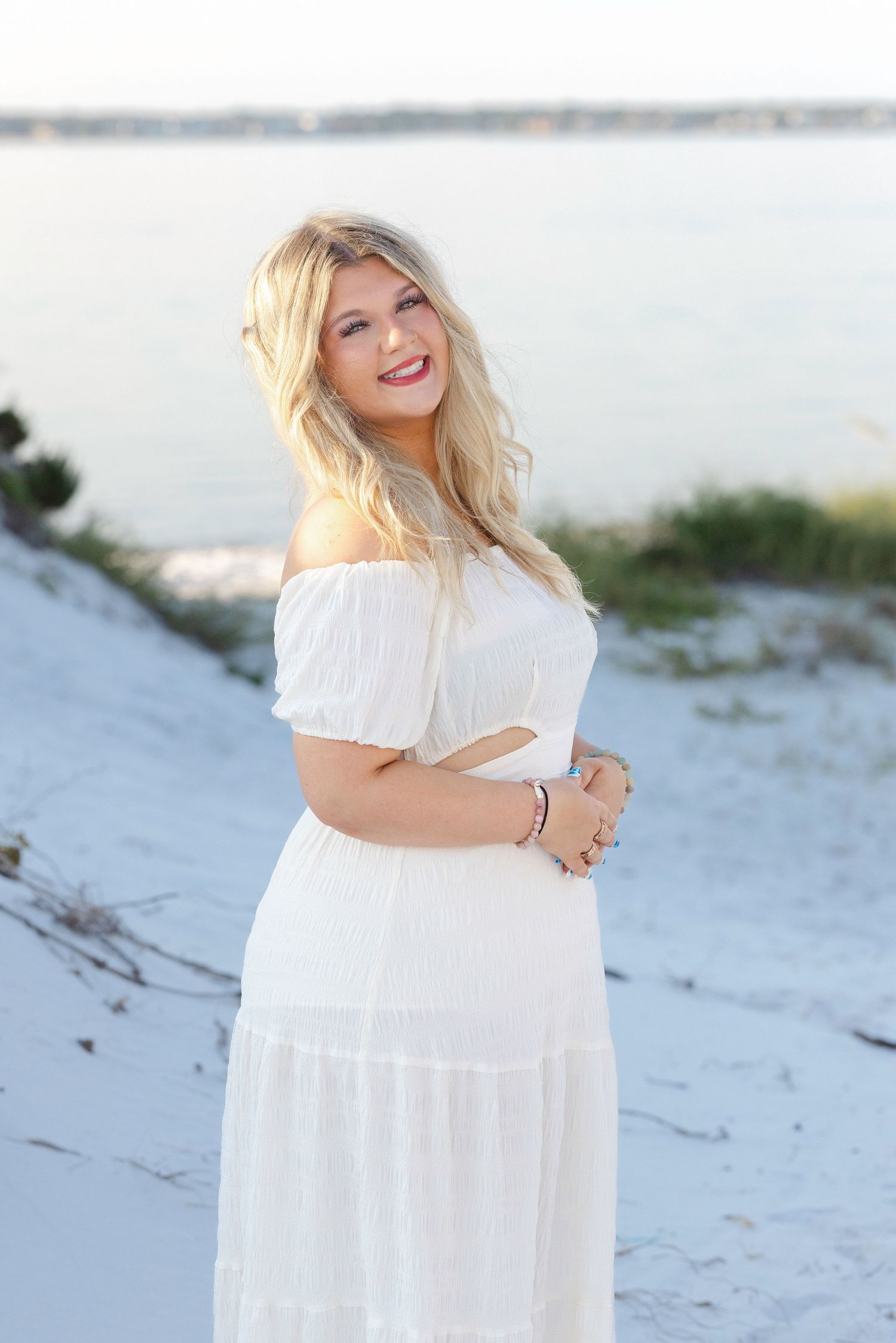 Smiling blonde woman in white off-shoulder maxi dress posing on sandy beach with water in background.