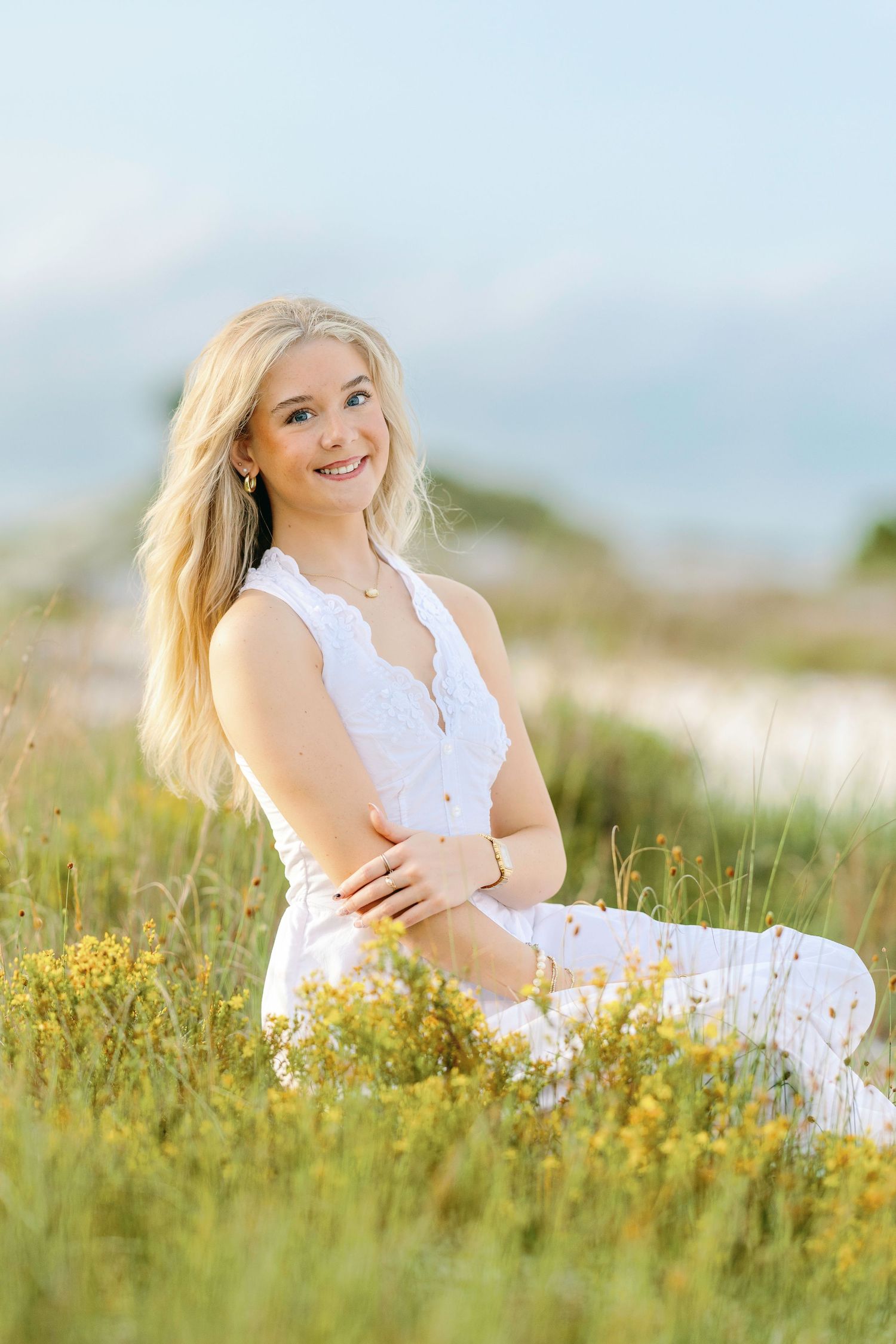 Smiling blonde teen in white dress sitting in wildflower field for senior portrait photo.