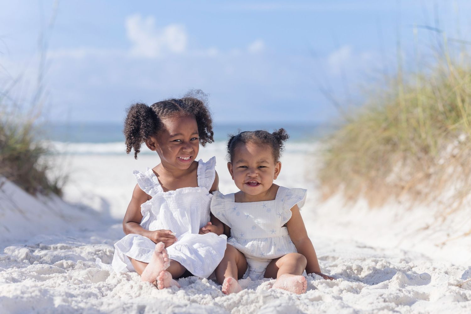 Two young sisters in white dresses smile while sitting on white sand at a sunny beach.