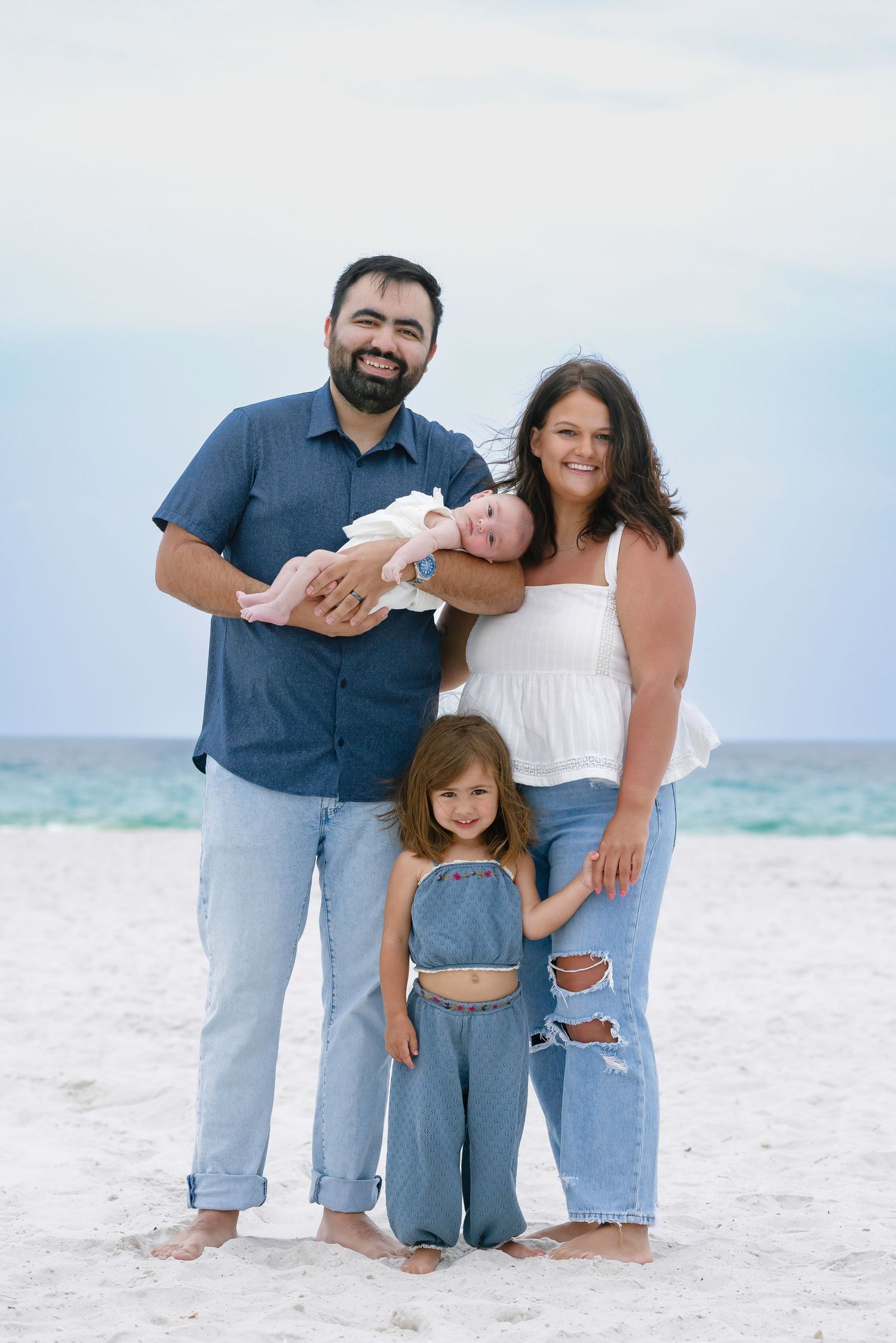 Family of four poses on white sand beach, parents holding newborn baby with toddler girl in denim overalls smiling.