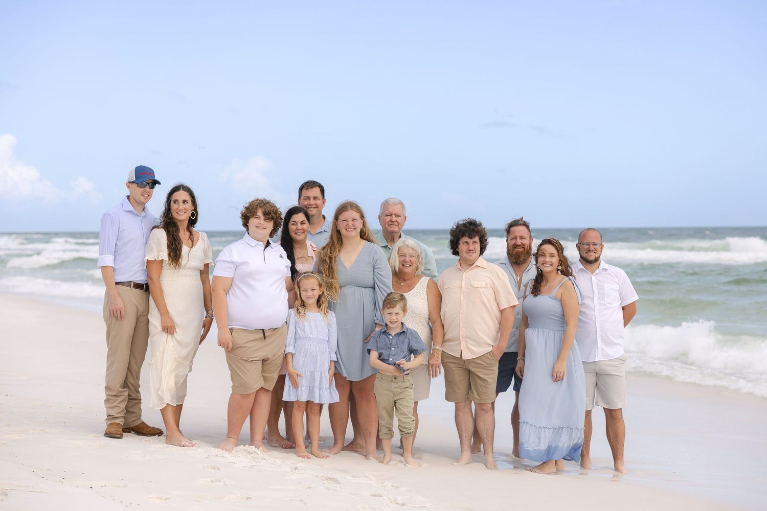 Large family gathering poses casually along beach shoreline wearing coordinated light colored clothing.