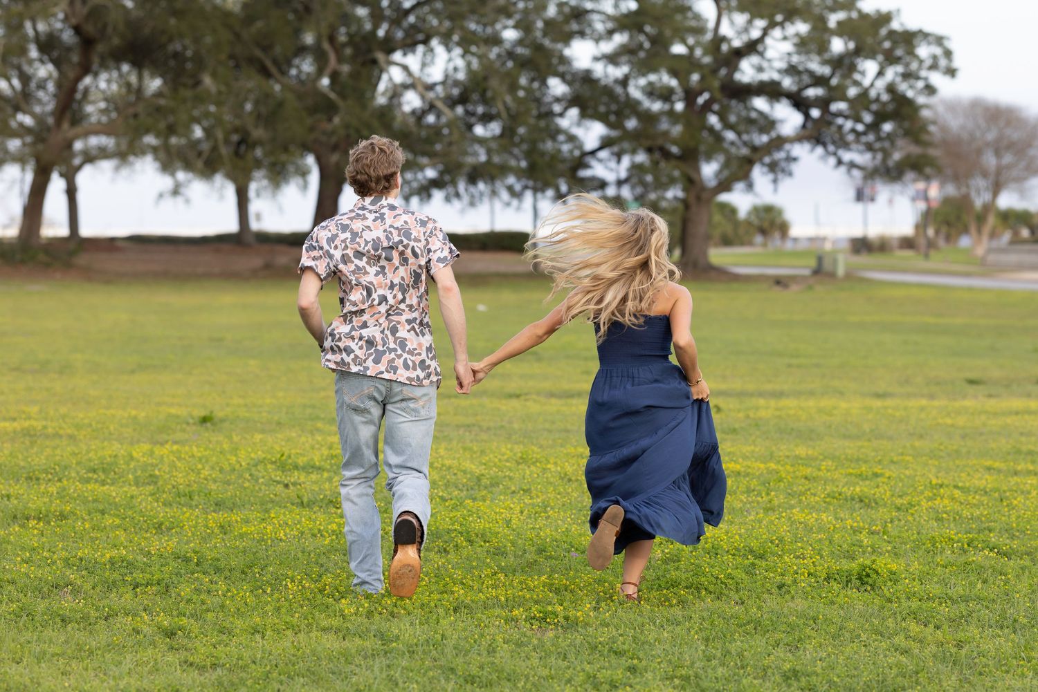 Two people run together holding hands across a green grass field with trees in the background.