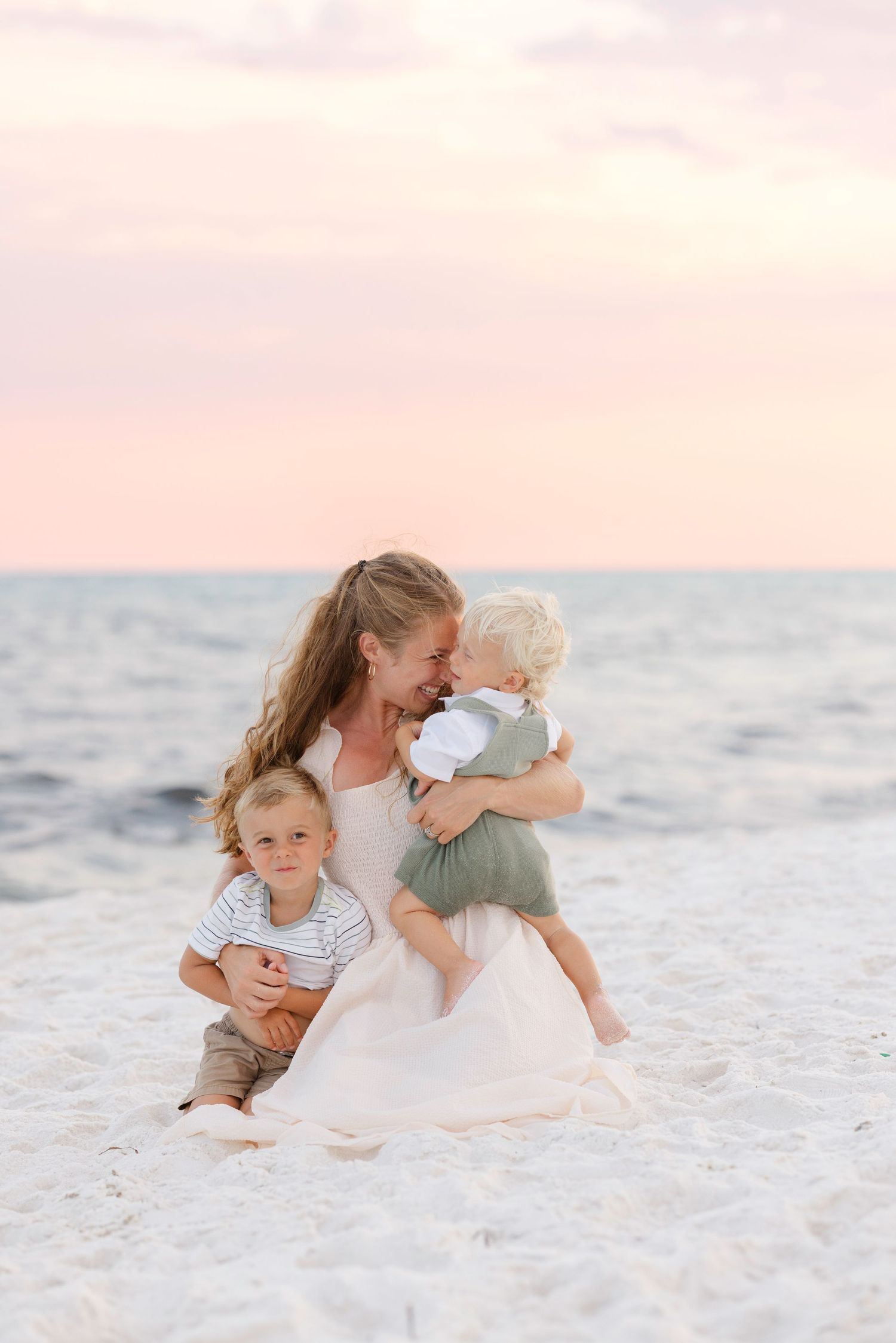 Family members embrace while sitting together on white sandy beach at sunset.