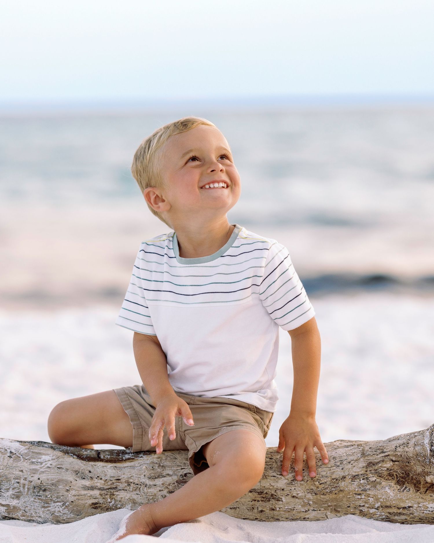 A child sits playfully on driftwood at the beach during a bright sunny day.