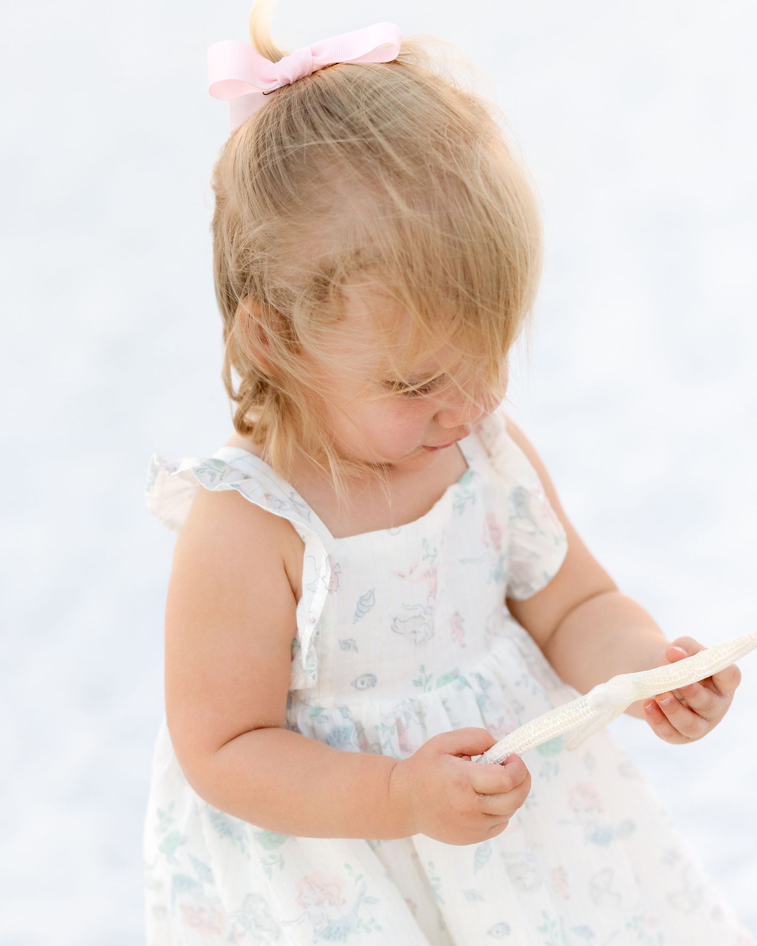 Adorable toddler girl in floral dress with pink bow examines small object at the beach.