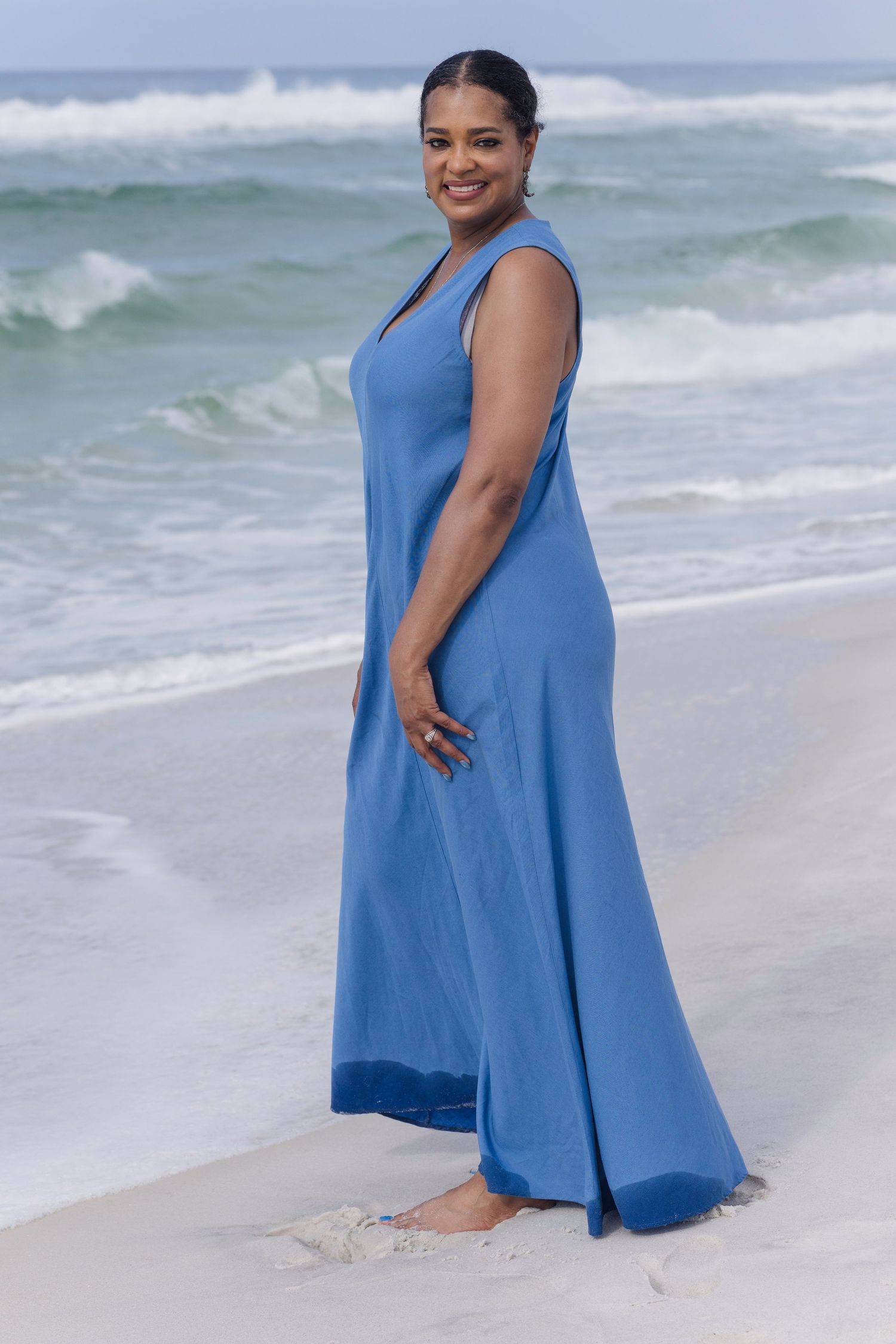 Woman in blue maxi dress smiling on sandy beach with ocean waves behind her.