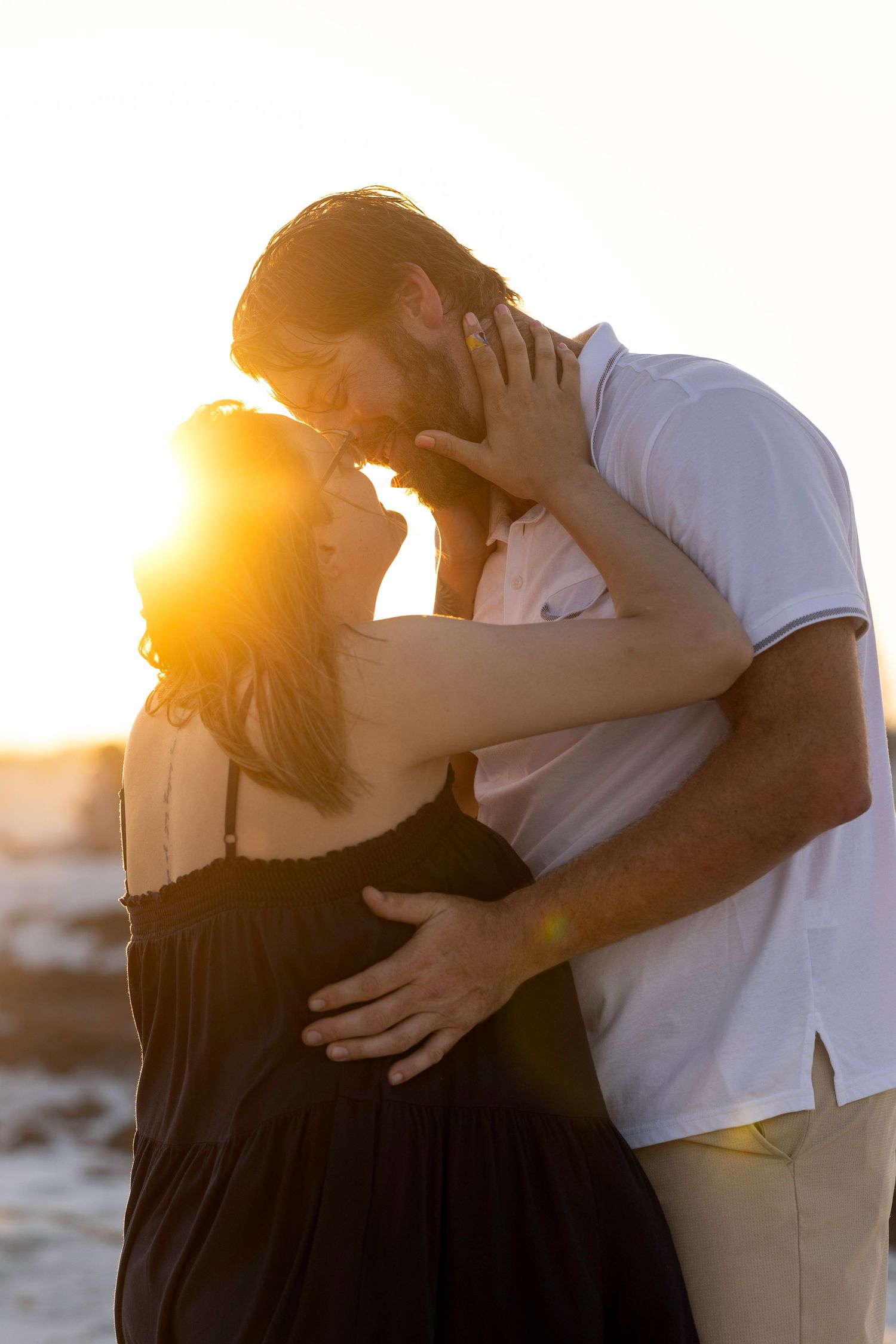 Romantic couple sharing a tender kiss at golden sunset on the beach, woman in black dress, man in white shirt.