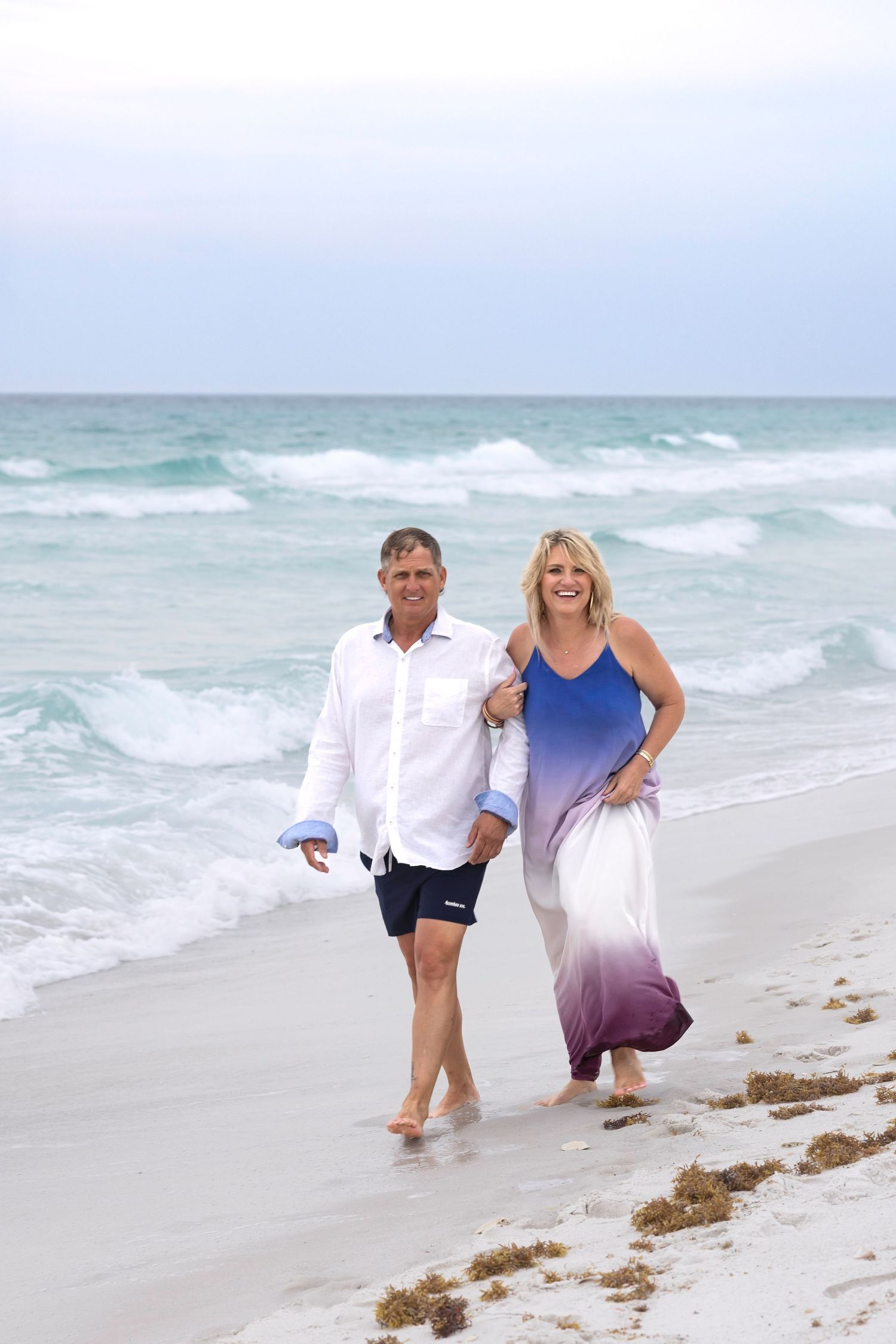 A smiling couple walks hand-in-hand along a sandy beach, with turquoise ocean waves in the background.