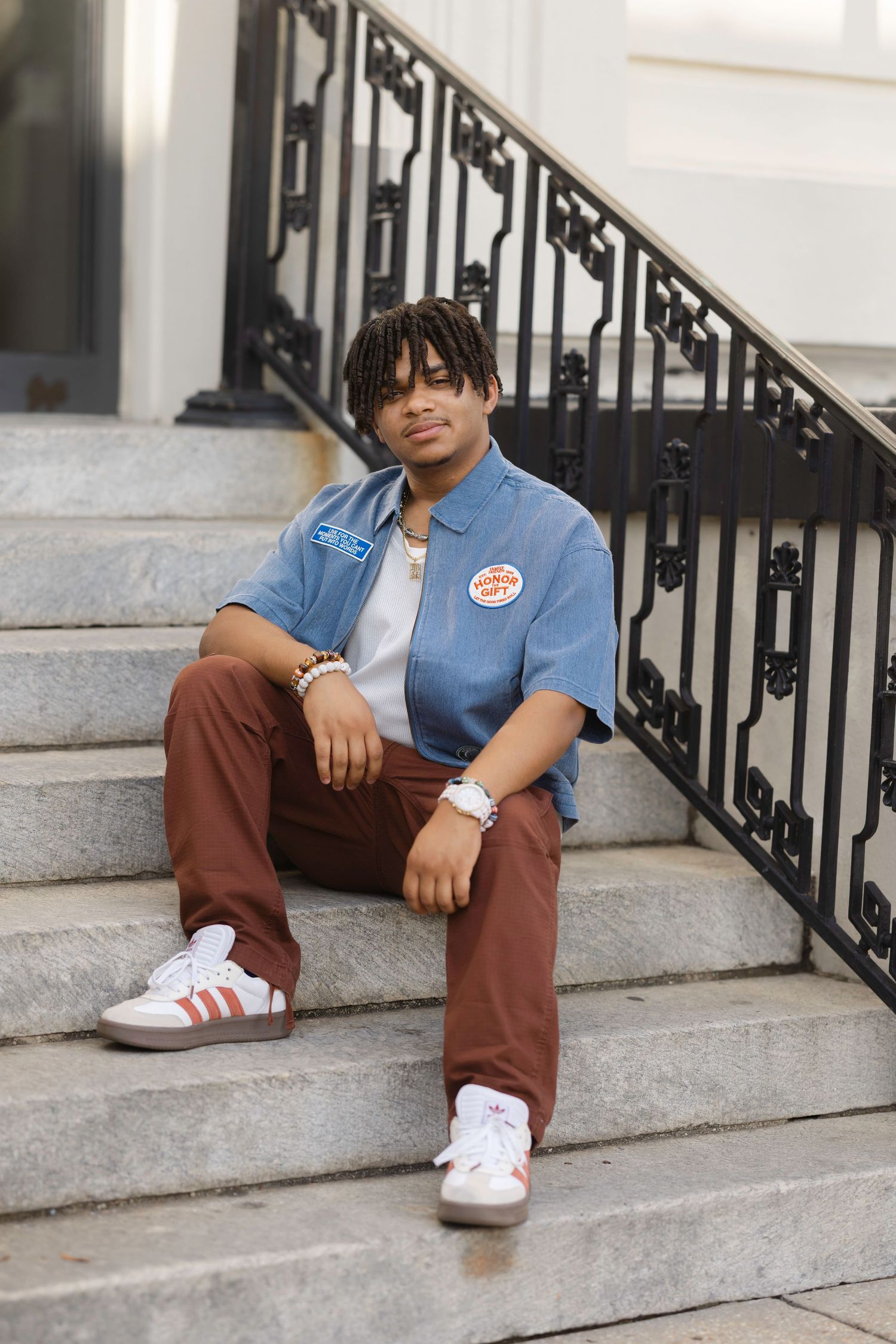 Young man with braids sits on stone steps wearing a blue shirt, brown pants, and white Adidas sneakers.