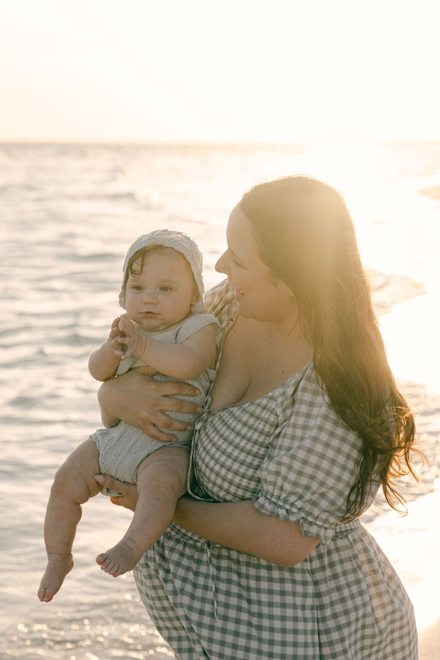 A mother holds her baby on a sunlit beach at golden hour, both wearing light outfits with the ocean glowing behind them.