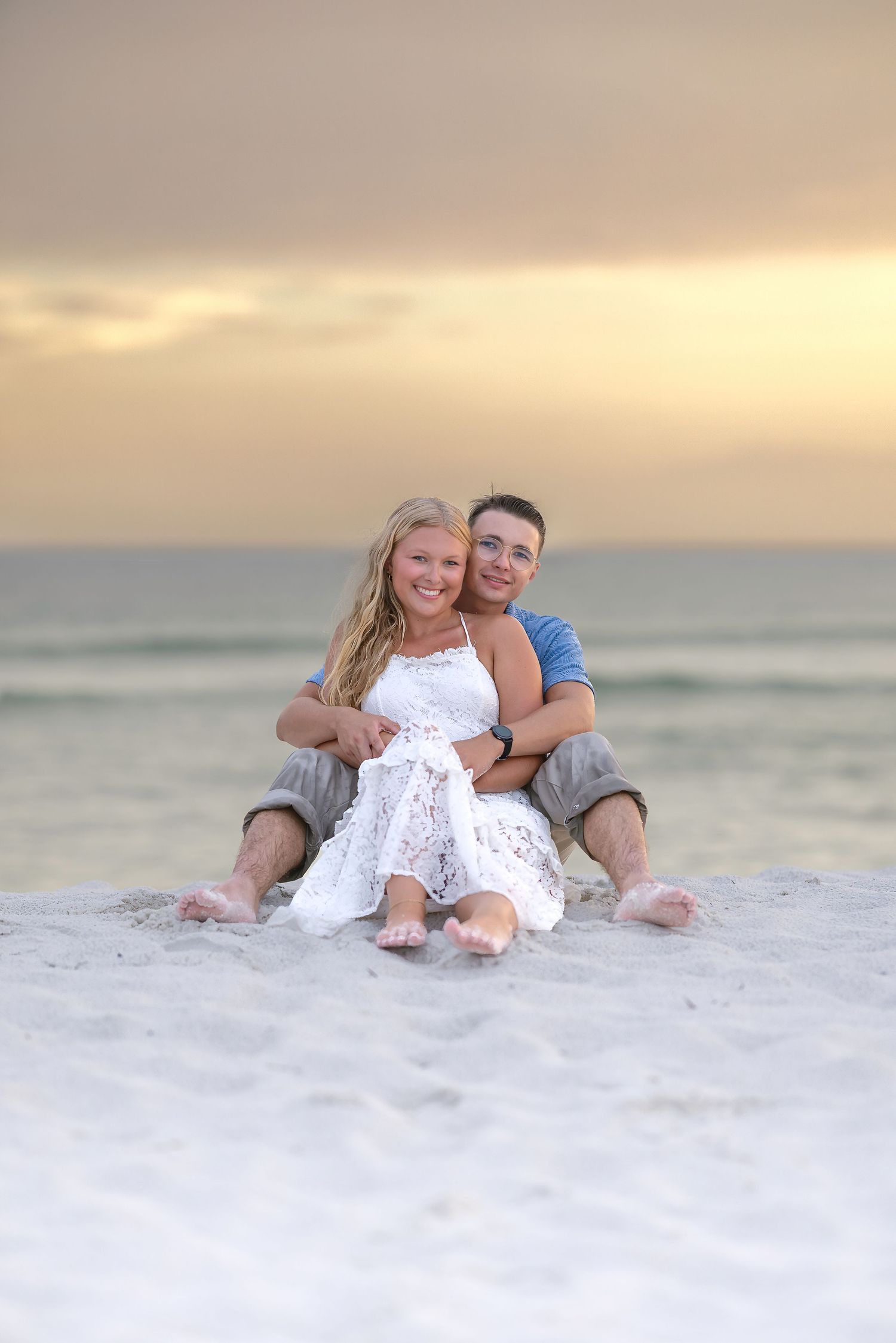 Couple sitting together on sandy beach at sunset, woman in white dress leaning back on man.