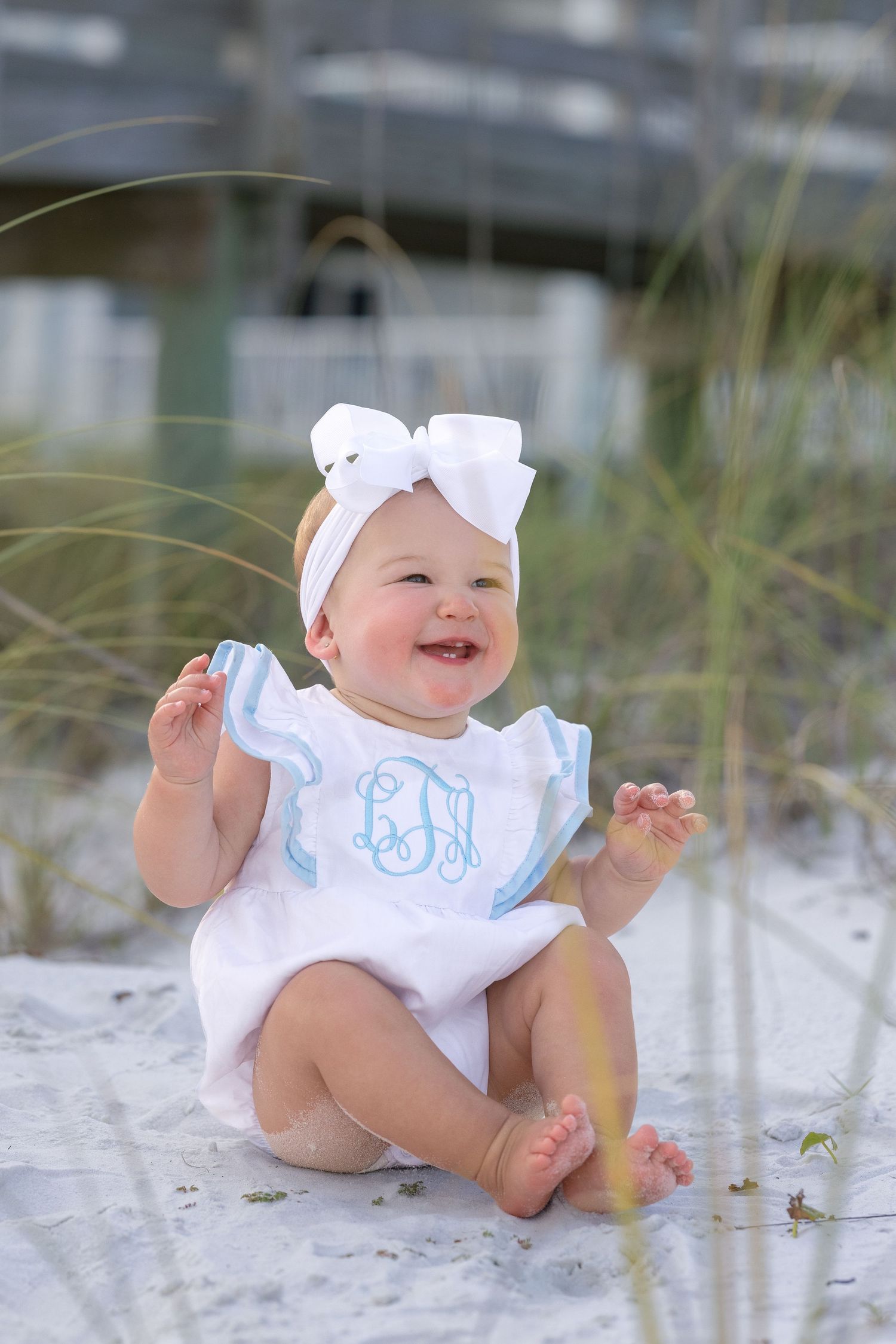 Happy baby girl in white monogrammed romper and bow headband sitting on white sand beach.