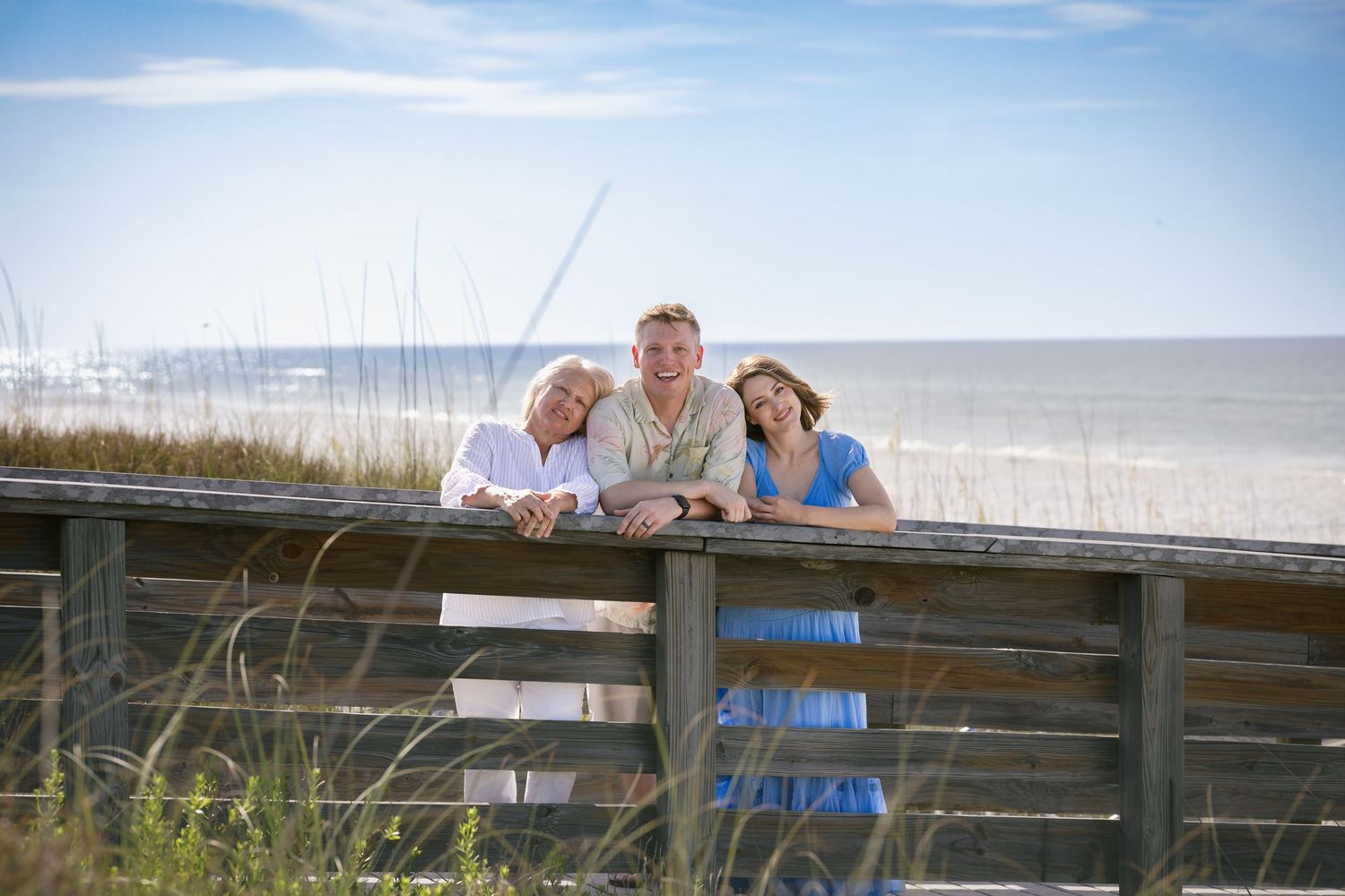 Three smiling people lean on a wooden boardwalk railing with a sandy beach and ocean in the background.