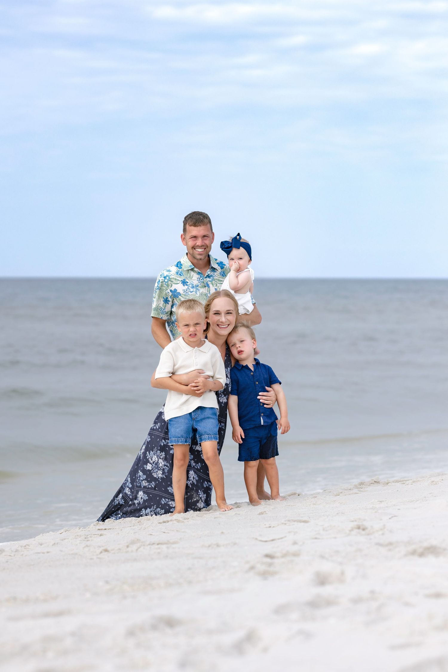 Happy family of five posing together on a sandy beach, with parents holding children near the ocean shoreline.