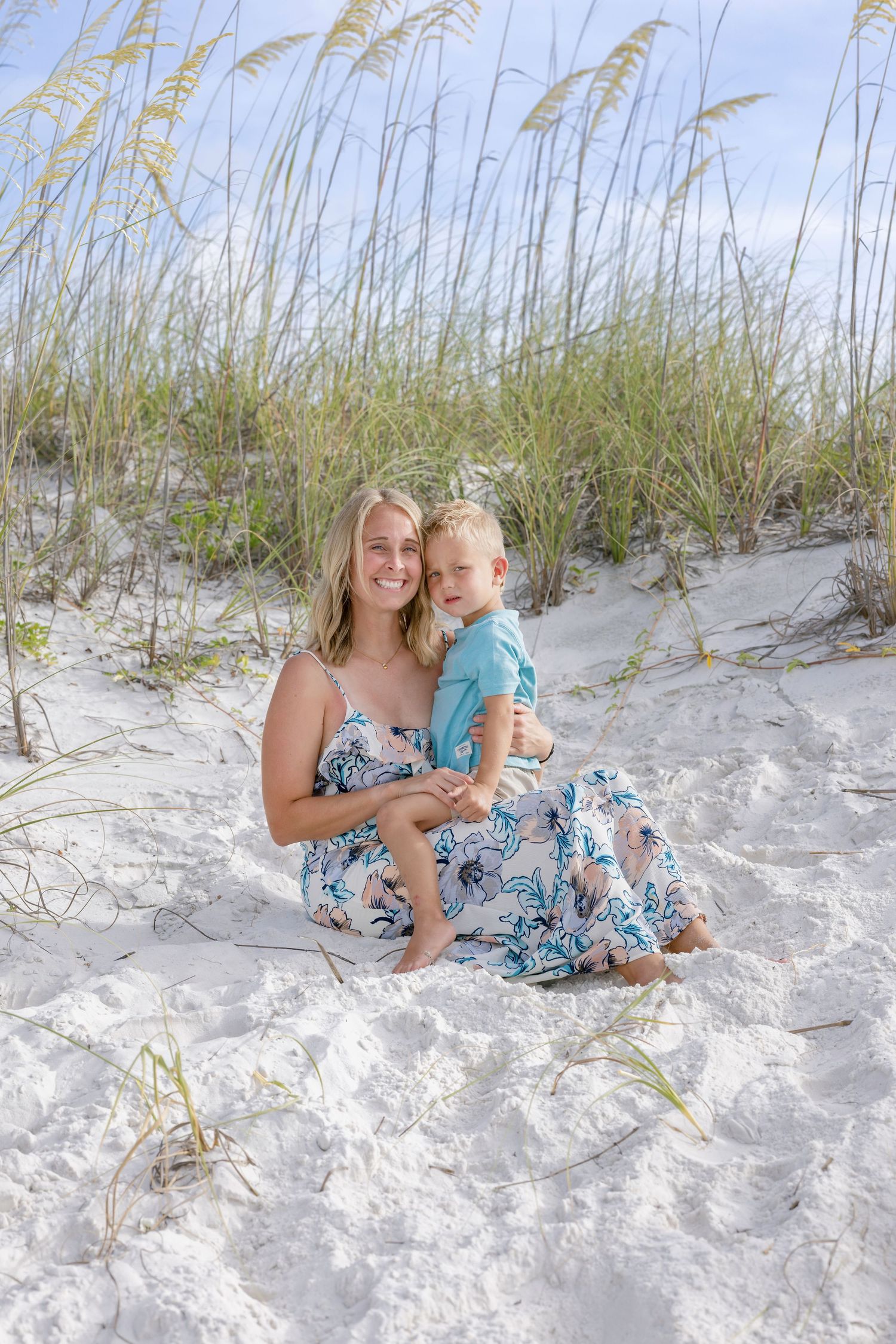 A smiling blonde mother sits on white sand dunes holding her toddler, both wearing blue floral outfits.
