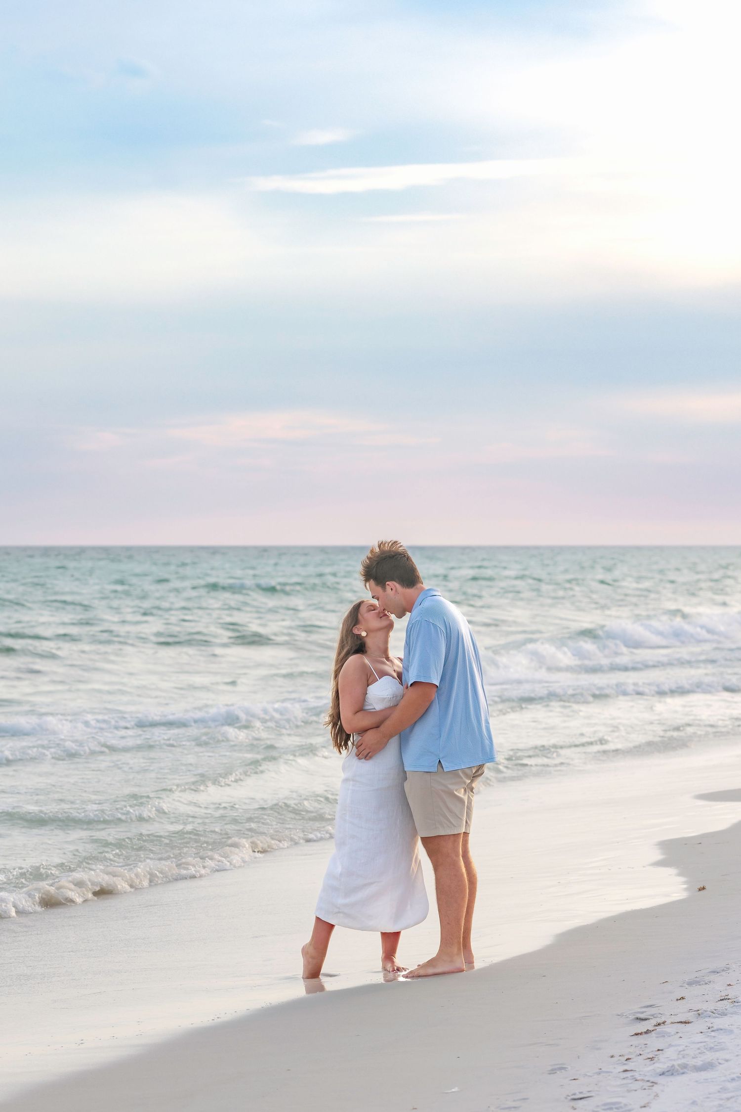 Romantic couple embracing and kissing on a white sand beach at sunset with turquoise ocean waves.