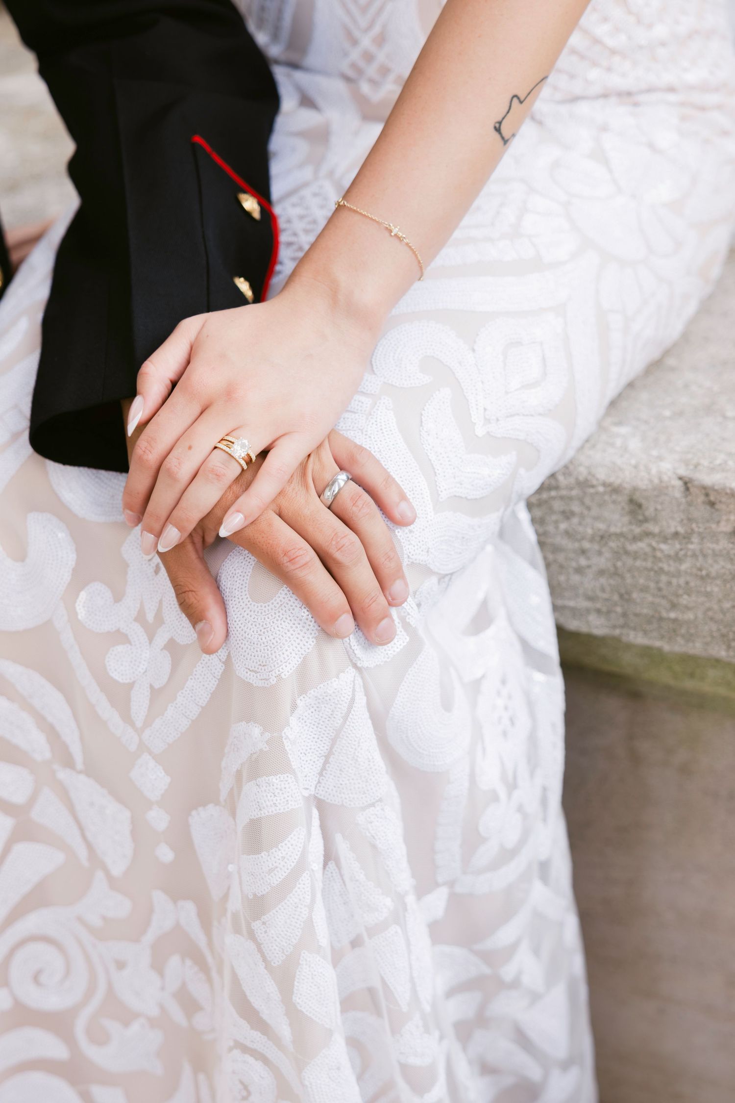 Newlywed couple displaying gold wedding rings, bride in white lace gown, groom in black military dress uniform.