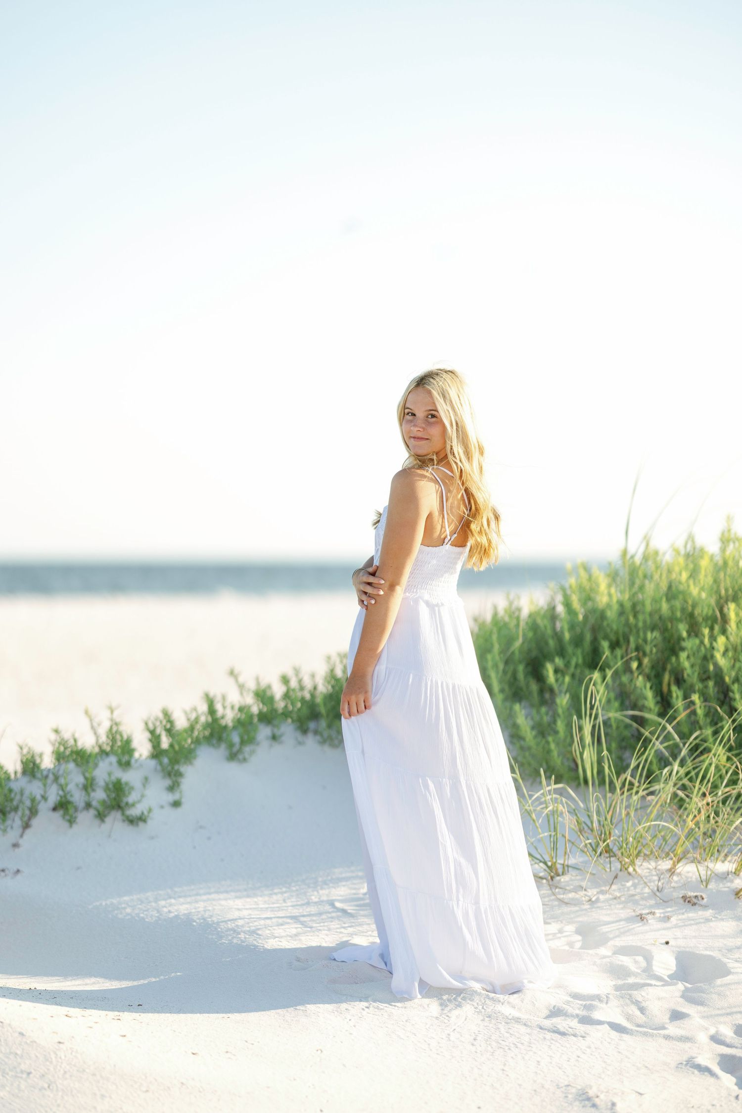 Woman in flowing white maxi dress poses gracefully on sandy beach dunes at sunset.