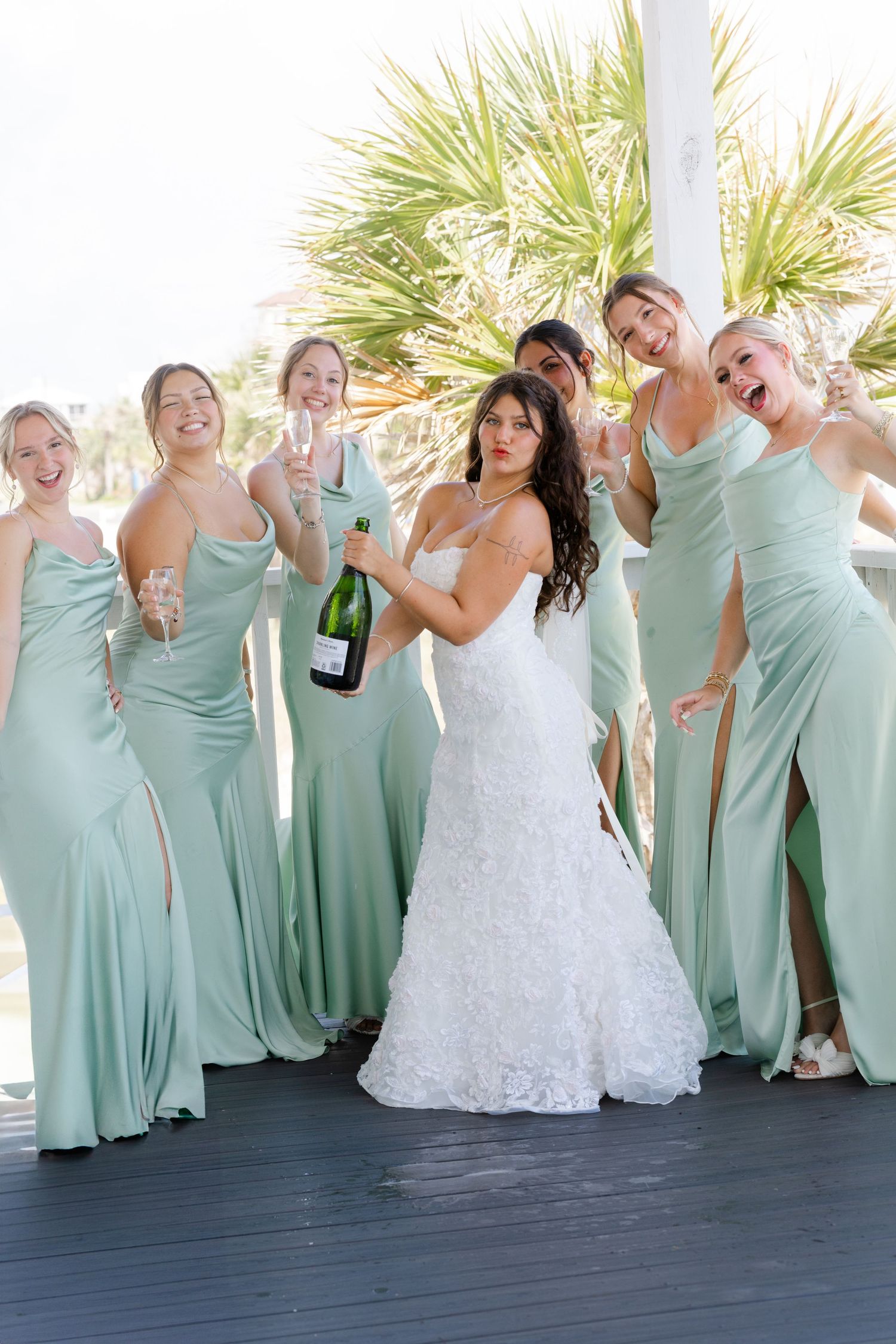 Bridesmaids in seafoam dresses celebrate with bride on beach deck with palm trees.